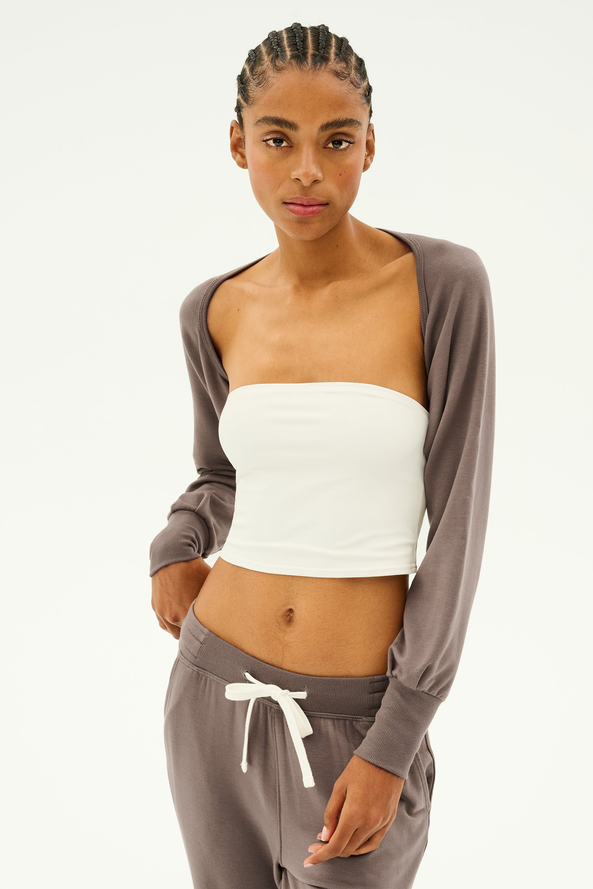 A person wearing the Sue Fleece Shrug in Lentil by SPLITS59, paired with a white tube top and matching pants, perfect for dance warmups, stands against a plain background.