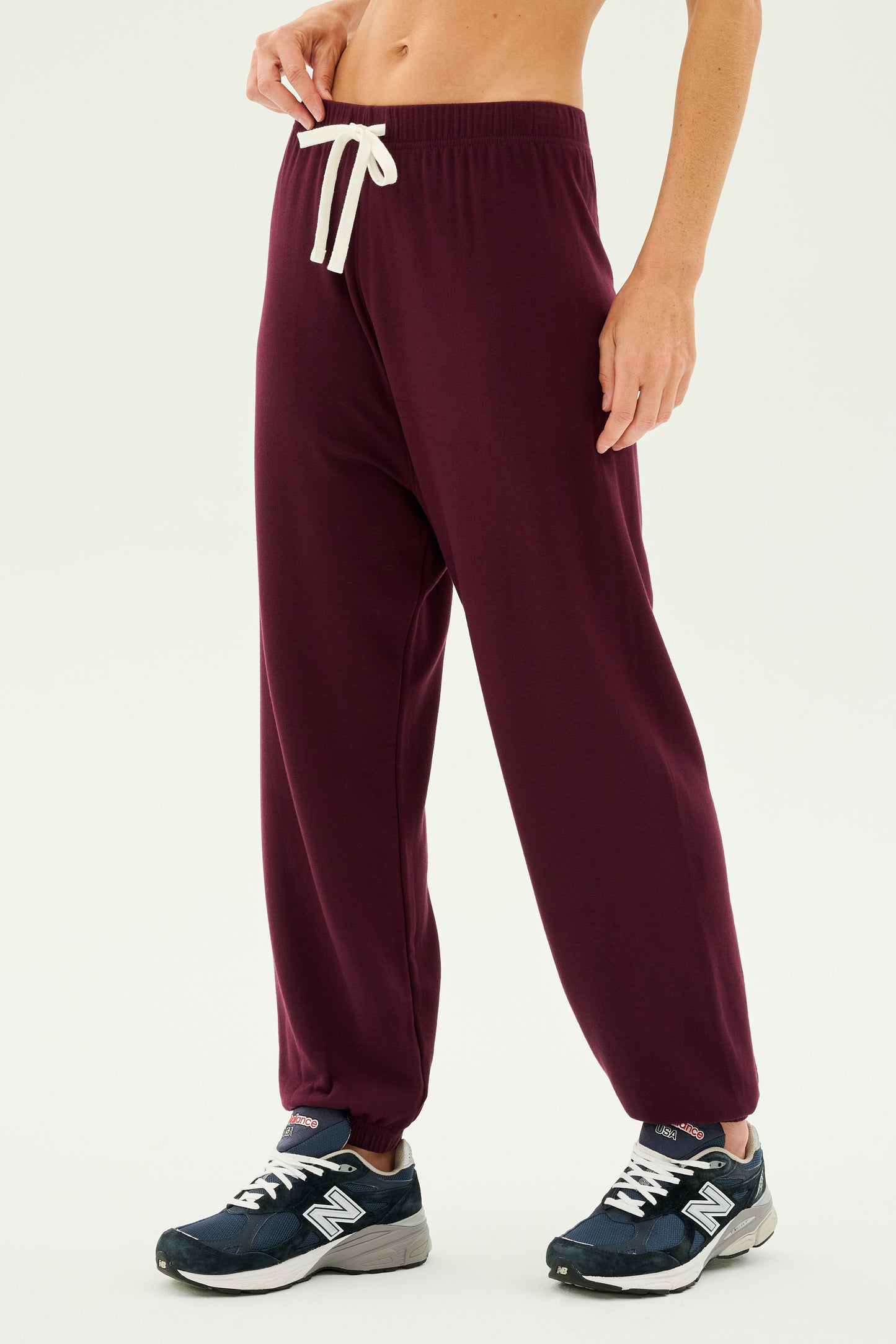 Person standing in Andie Oversized Fleece Sweatpant - Dark Cherry by SPLITS59 with a white drawstring and black running shoes with white details against a plain white background, ready for cool weather workouts.