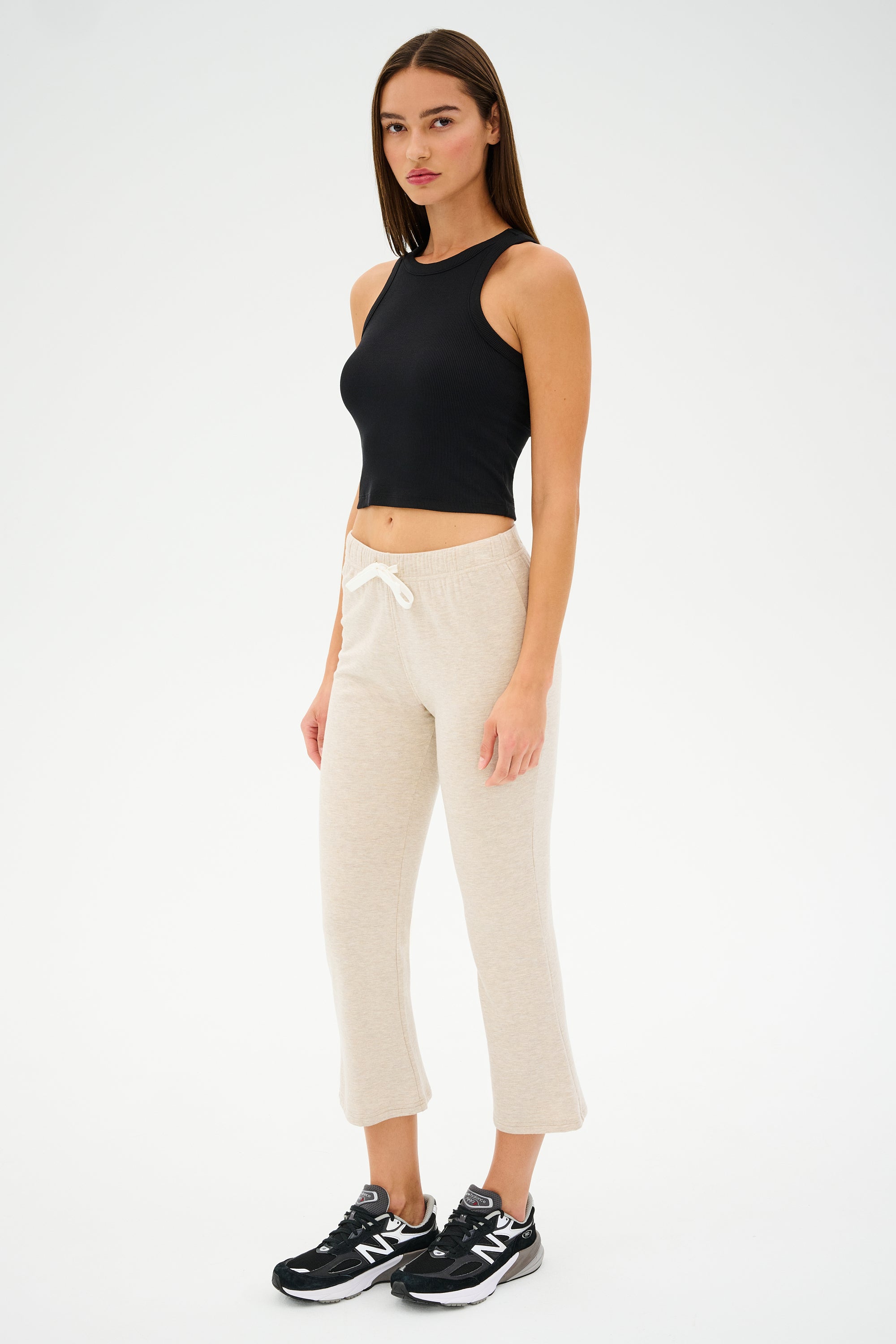 A woman in a black sleeveless top, Brooks Fleece Cropped Flare - Heather Oatmeal pants from SPLITS59, and black New Balance sneakers stands against a plain white background, embodying a comfy casual chic style.