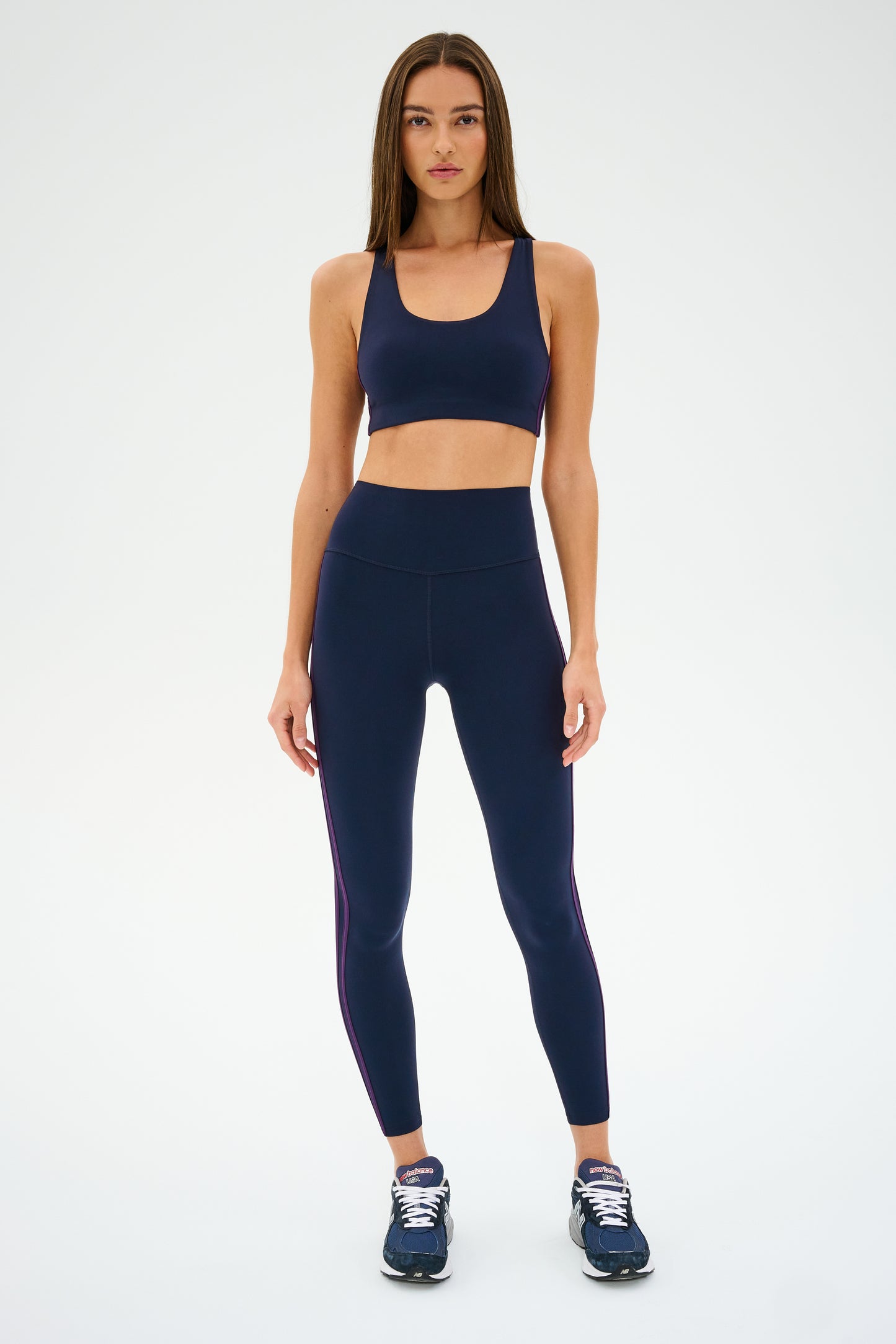 A woman poses in the Ella Airweight Bra by SPLITS59, navy blue sportswear leggings with side stripes, and sneakers against a plain backdrop.