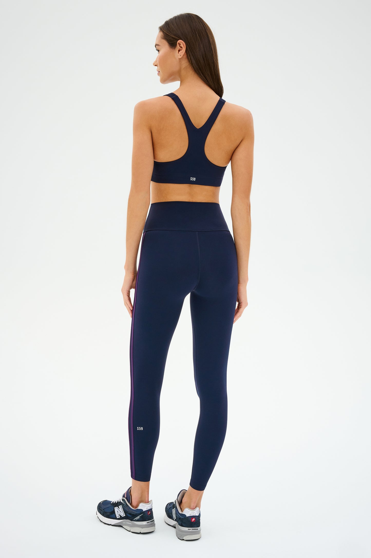 A woman in navy athletic wear, including the SPLITS59 Ella Airweight Bra in Indigo/Black Currant and sneakers, stands facing away on a white background. The Airweight fabric is ideal for hot yoga sessions.