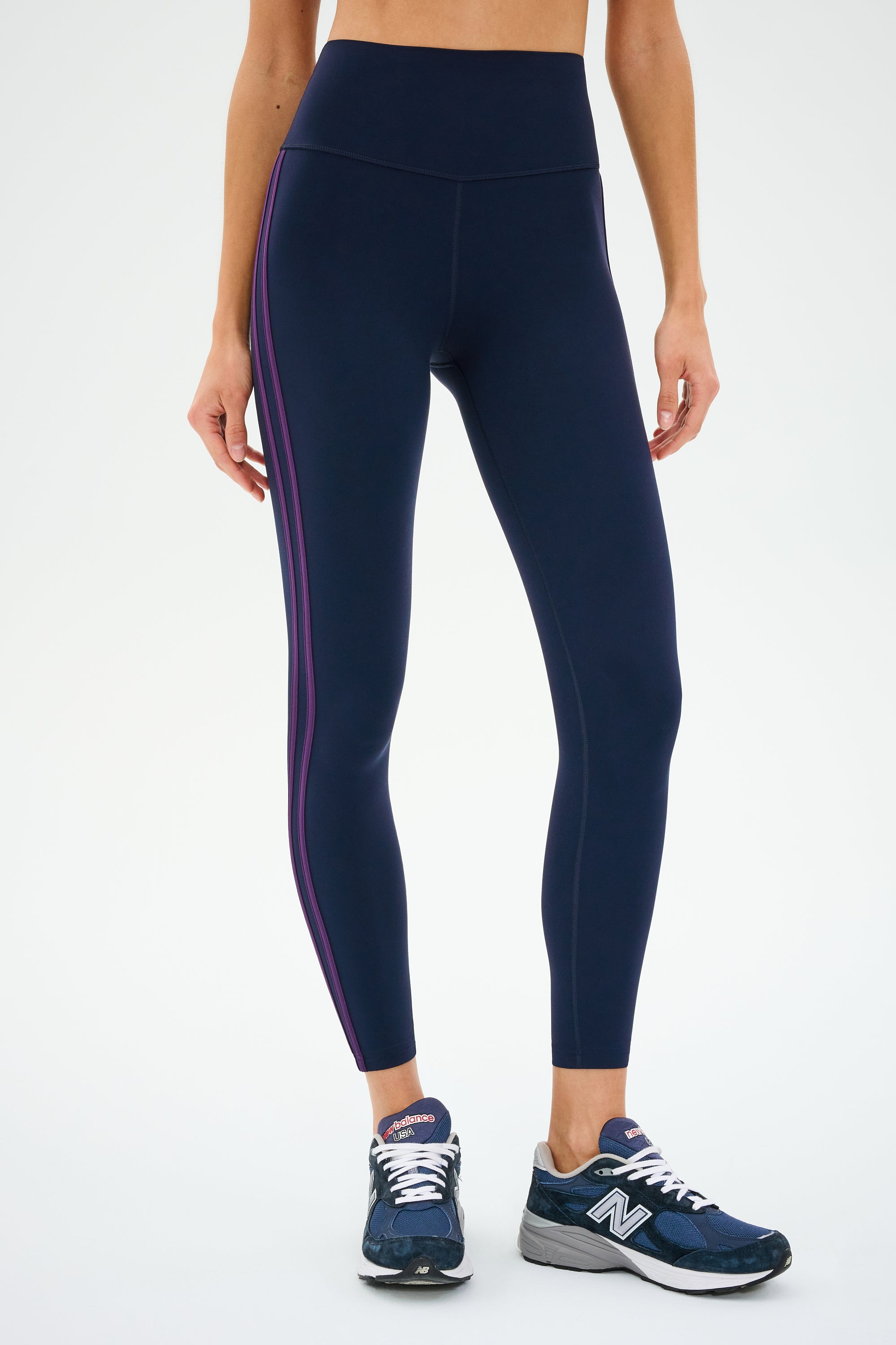 Someone in SPLITS59's Ella High Waist Airweight 7/8 leggings in Indigo/Black Currant and navy sneakers stands against a plain backdrop. The butter-soft fabric is perfect for hot yoga, though their head isn't visible.