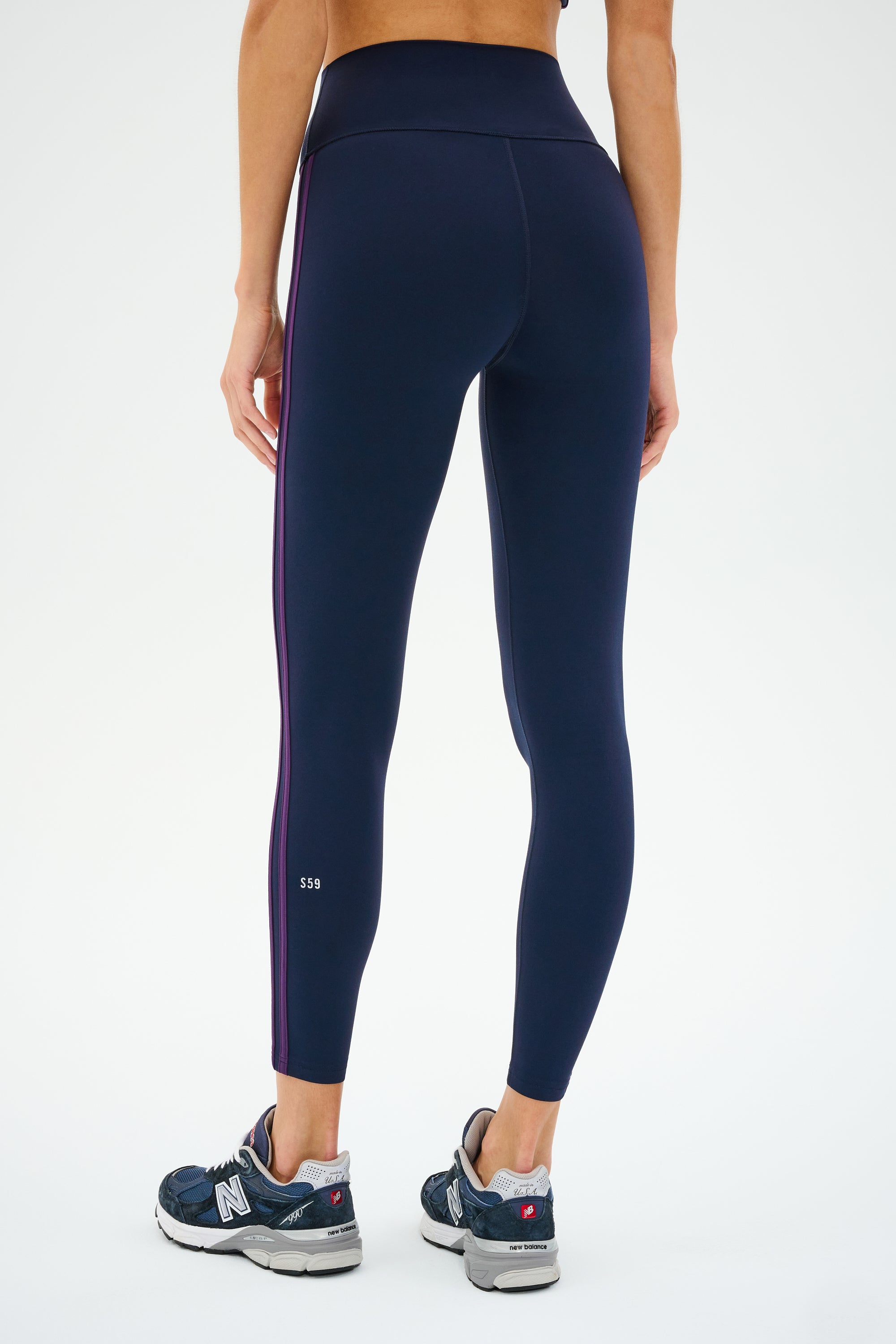 A person wearing SPLITS59's Ella High Waist Airweight 7/8 leggings in Indigo/Black Currant and blue athletic shoes stands on a white background, viewed from the back.