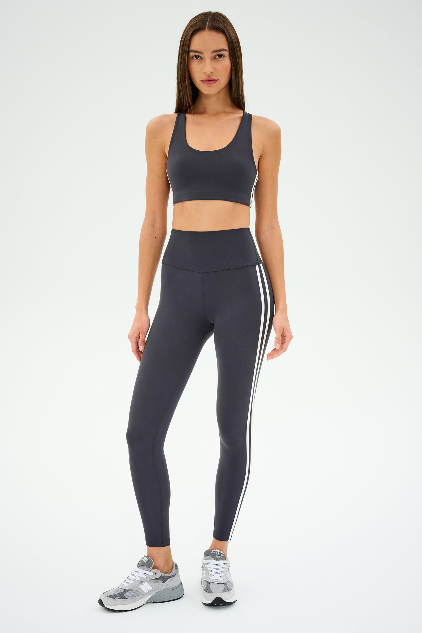 A person stands against a white background wearing SPLITS59 Ella High Waist Airweight 7/8 leggings in Graphite/White with a matching sports bra and gray sneakers.