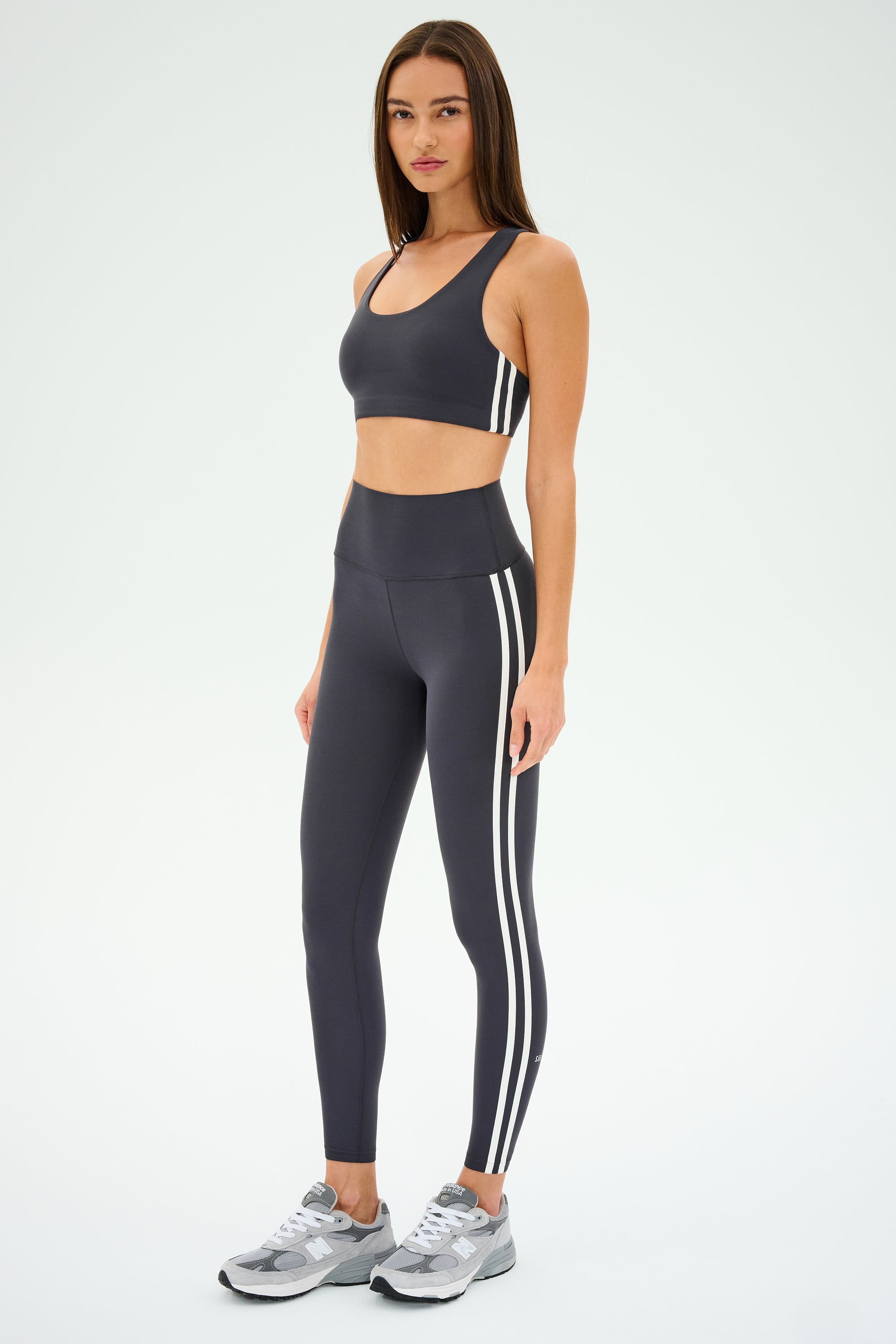 A person wearing SPLITS59's Ella High Waist Airweight 7/8 leggings in Graphite/White and a gray sports bra, paired with gray sneakers, stands against a plain background.