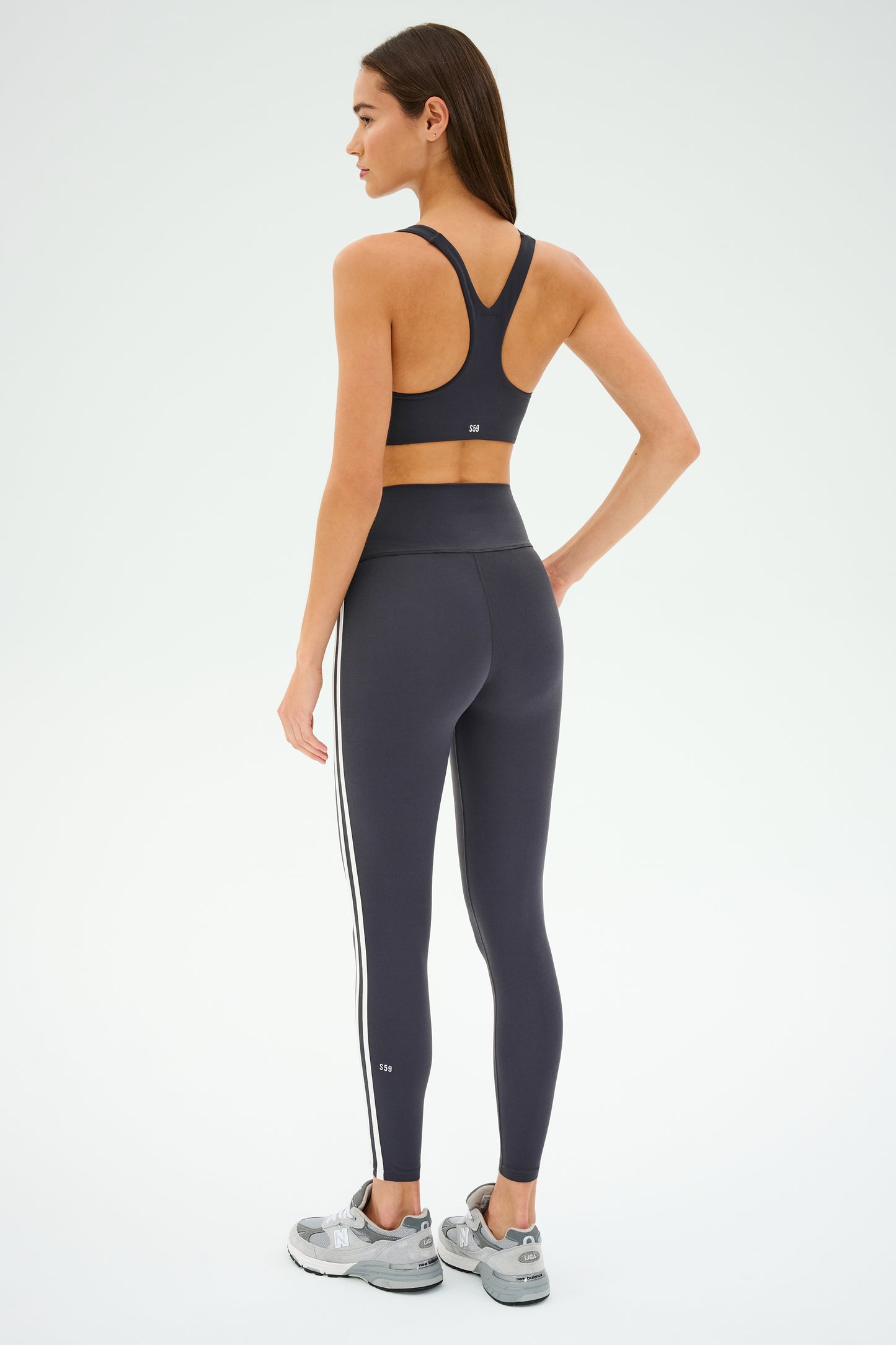 A woman stands with her back to the camera, wearing SPLITS59's Ella High Waist Airweight 7/8 in Graphite/White. She's in a sports bra, sneakers, and has long straight hair, ready for her hot yoga session.