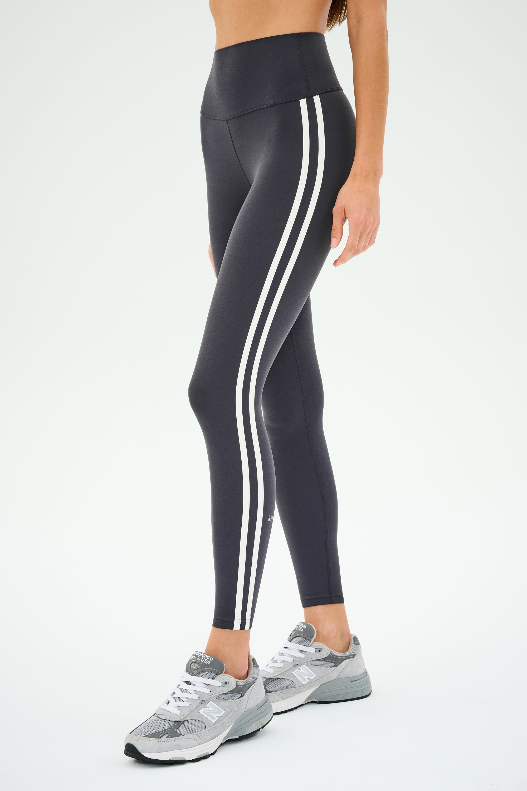 An individual is wearing SPLITS59's Ella High Waist Airweight 7/8 leggings in graphite with white stripes, paired with gray sneakers, standing against a plain background.