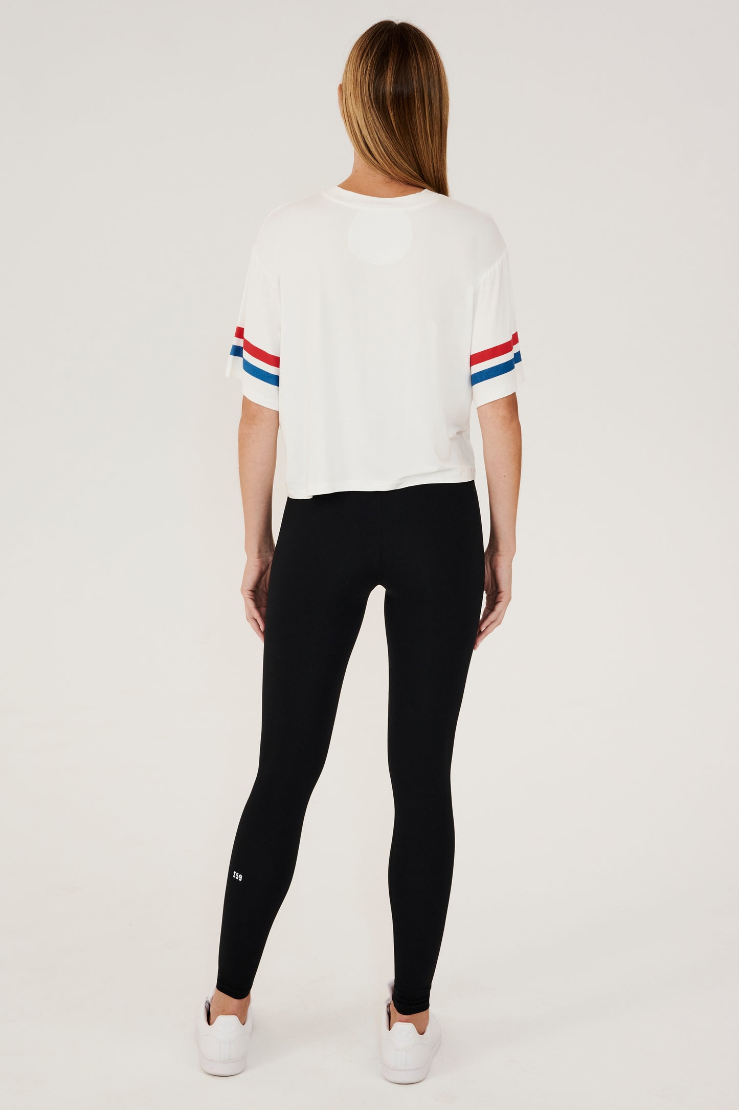 Full back view of girl wearing a short sleeve cropped white shirt with red and blue stripes on the arm and a black leggings with white shoes