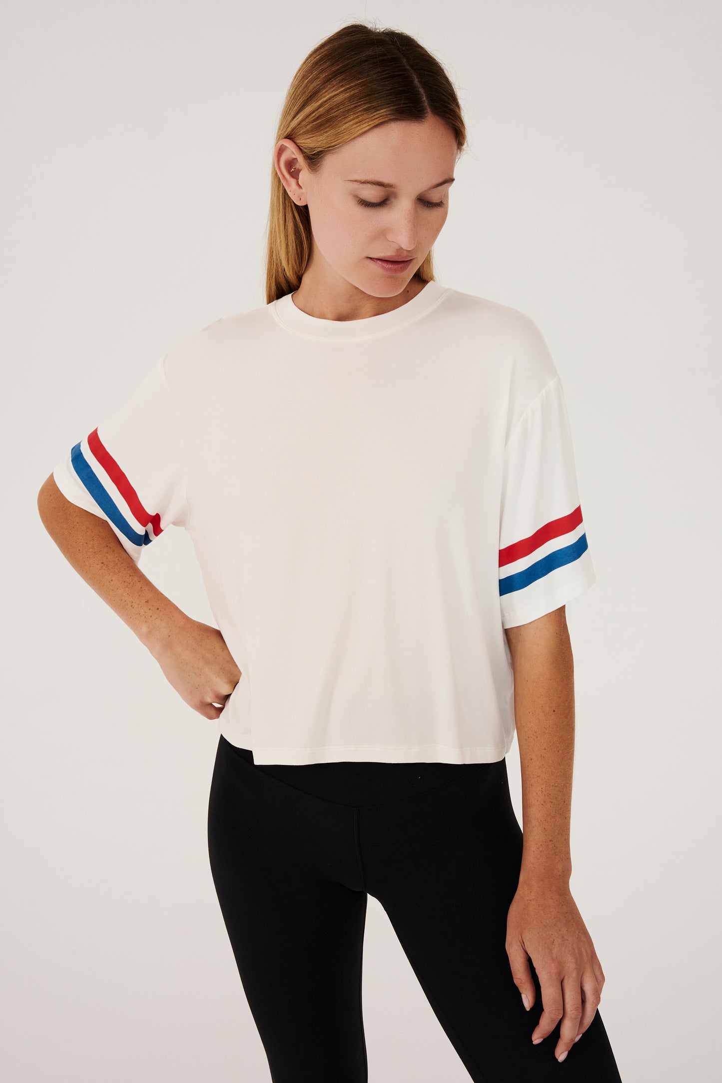 Front view of girl wearing a short sleeve cropped white shirt with red and blue stripes on the arm and a black leggings 