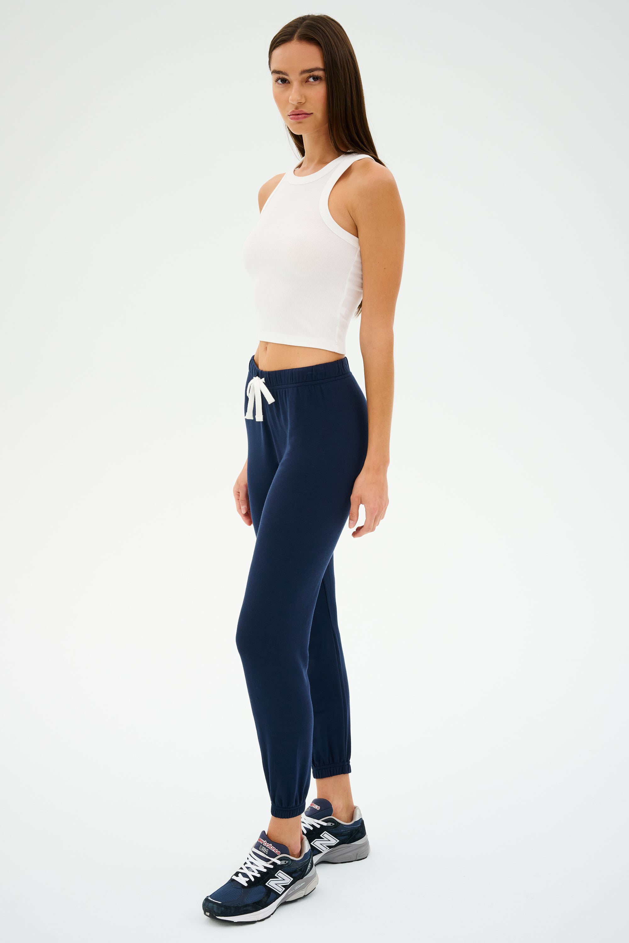 A person stands wearing a white sleeveless top, SPLITS59 Sonja Fleece Sweatpant in Indigo, and sneakers, posing sideways against a plain white background.