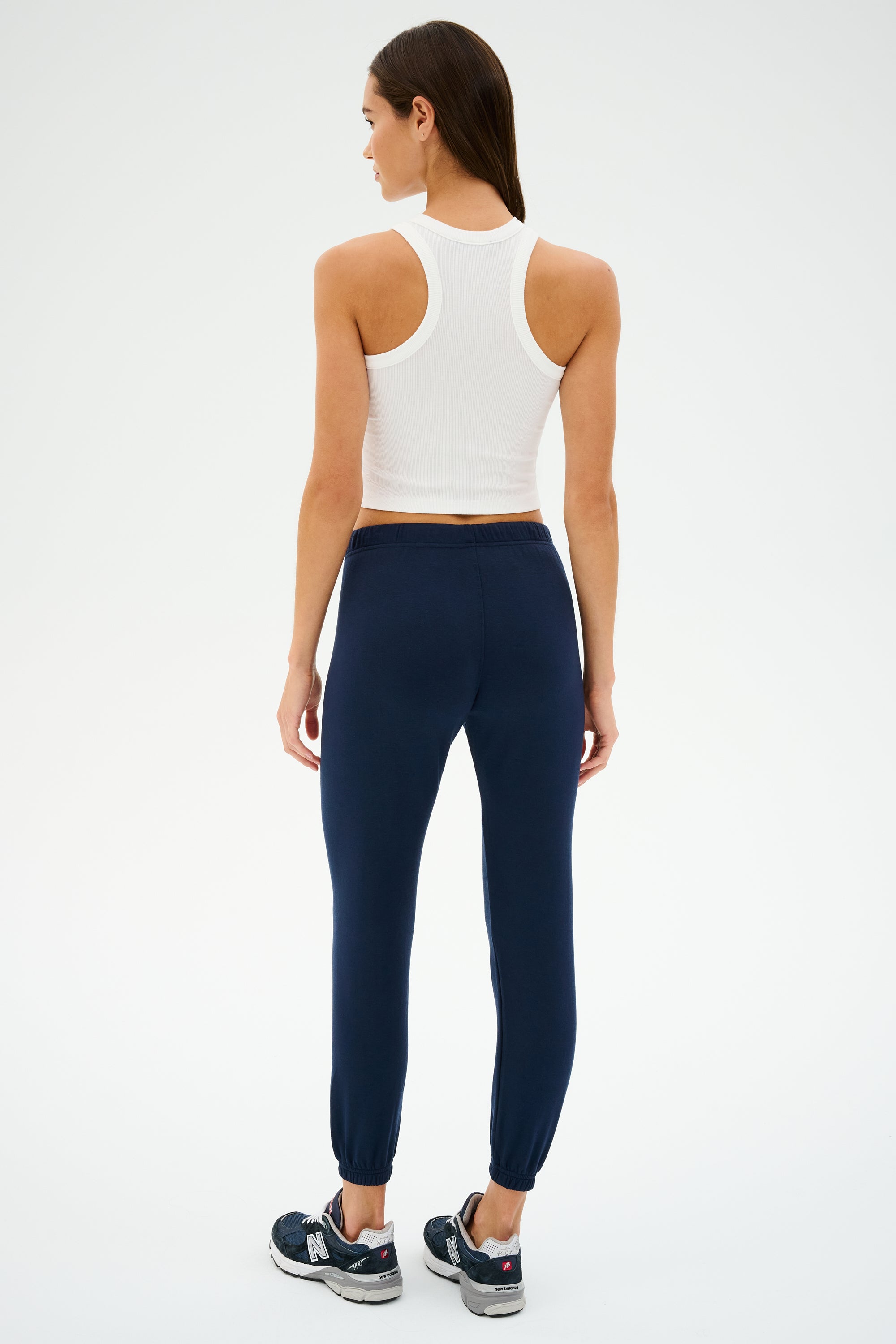 Facing away from the camera, this person models a white tank top and SPLITS59's Sonja Fleece Sweatpant in Indigo, ideal for cool-weather workouts. The combination of navy joggers with black sneakers creates a sleek profile against a plain white backdrop.