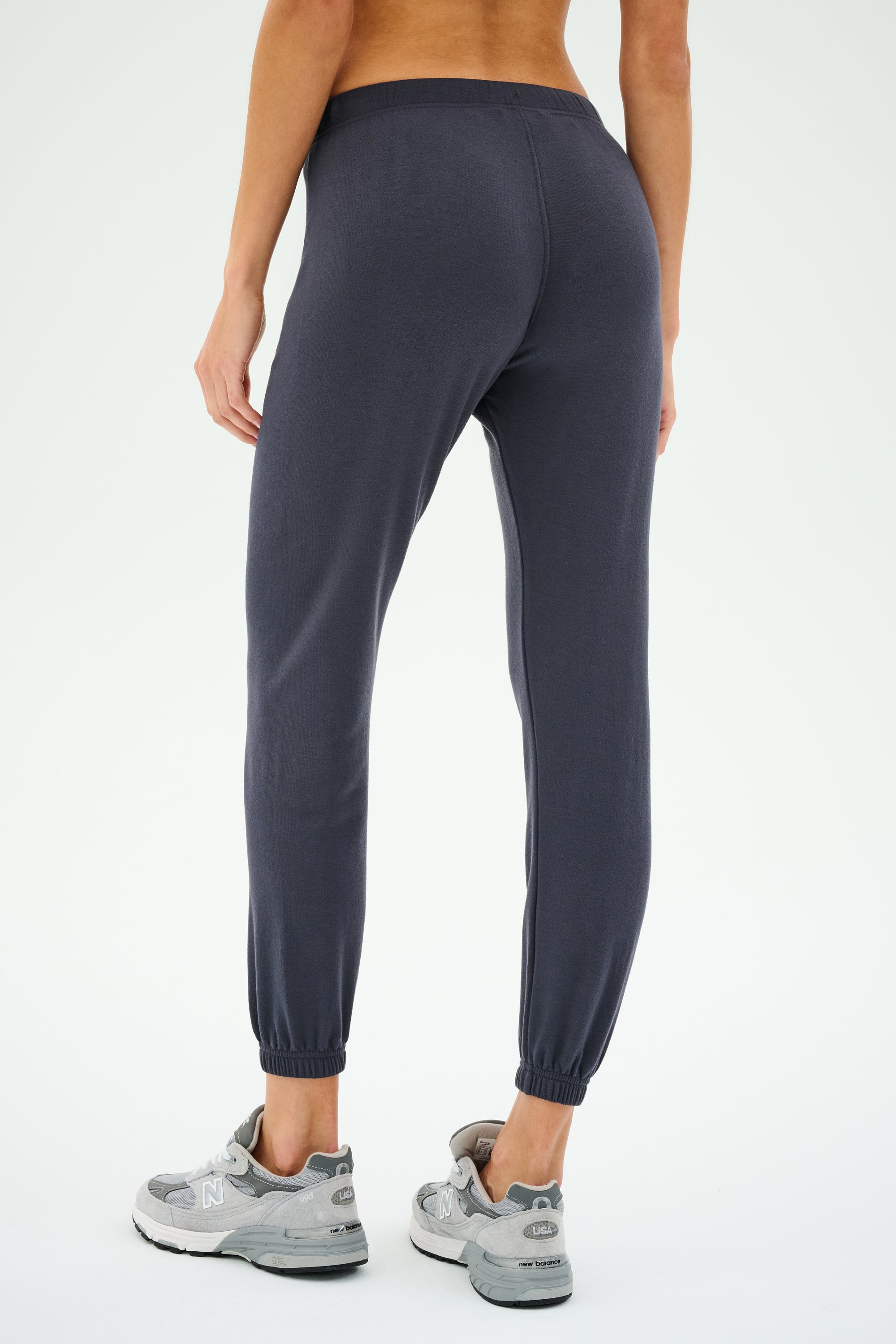 A person wearing SPLITS59's Sonja Fleece Sweatpant in Graphite and gray sneakers stands with their back turned. Ideal for cool weather workouts, these slouchy-soft pants offer unmatched comfort and style.