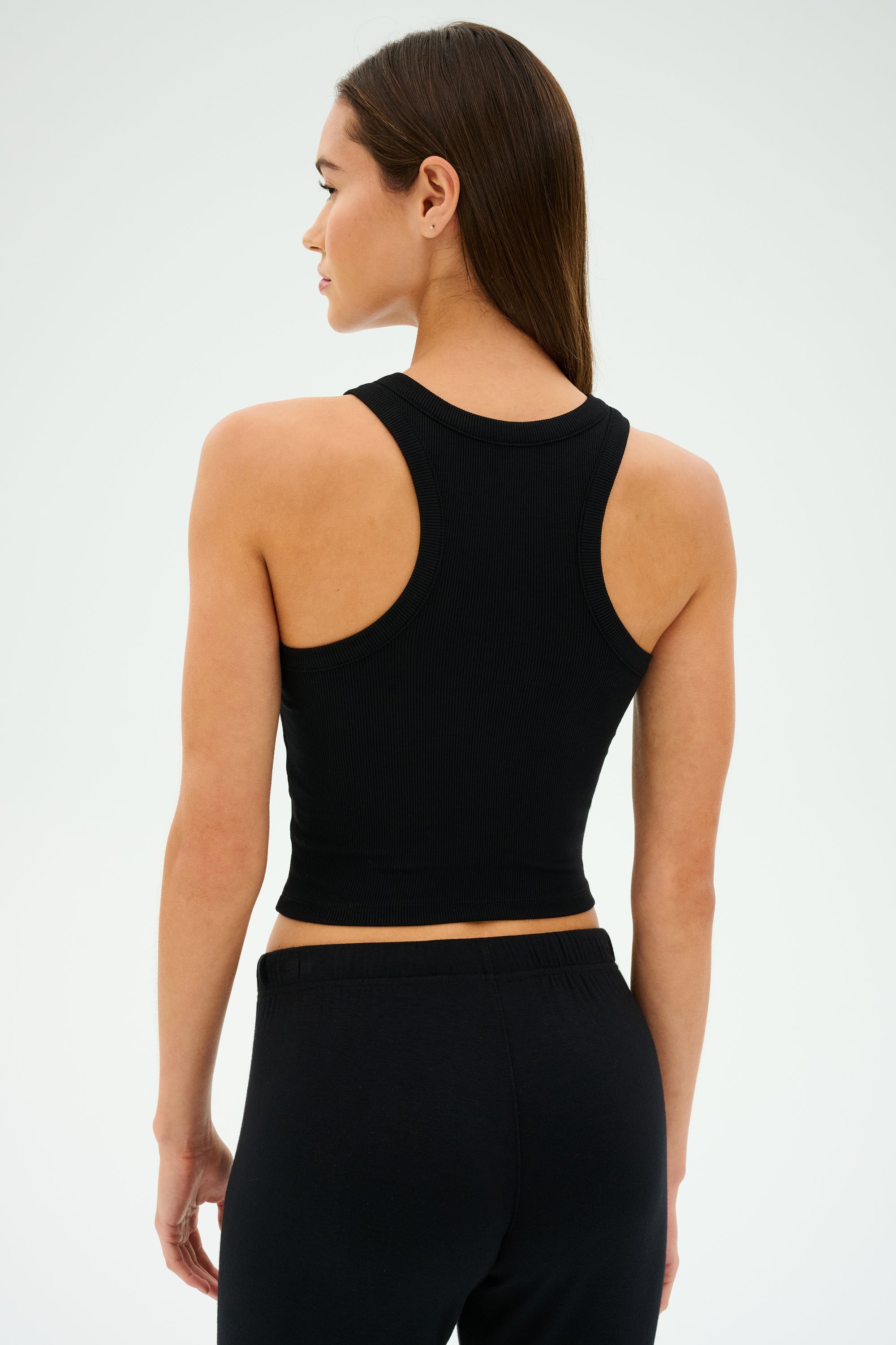 A person with long brown hair is wearing the SPLITS59 Kiki Rib Crop Tank in black and black pants, facing sideways against a plain background.