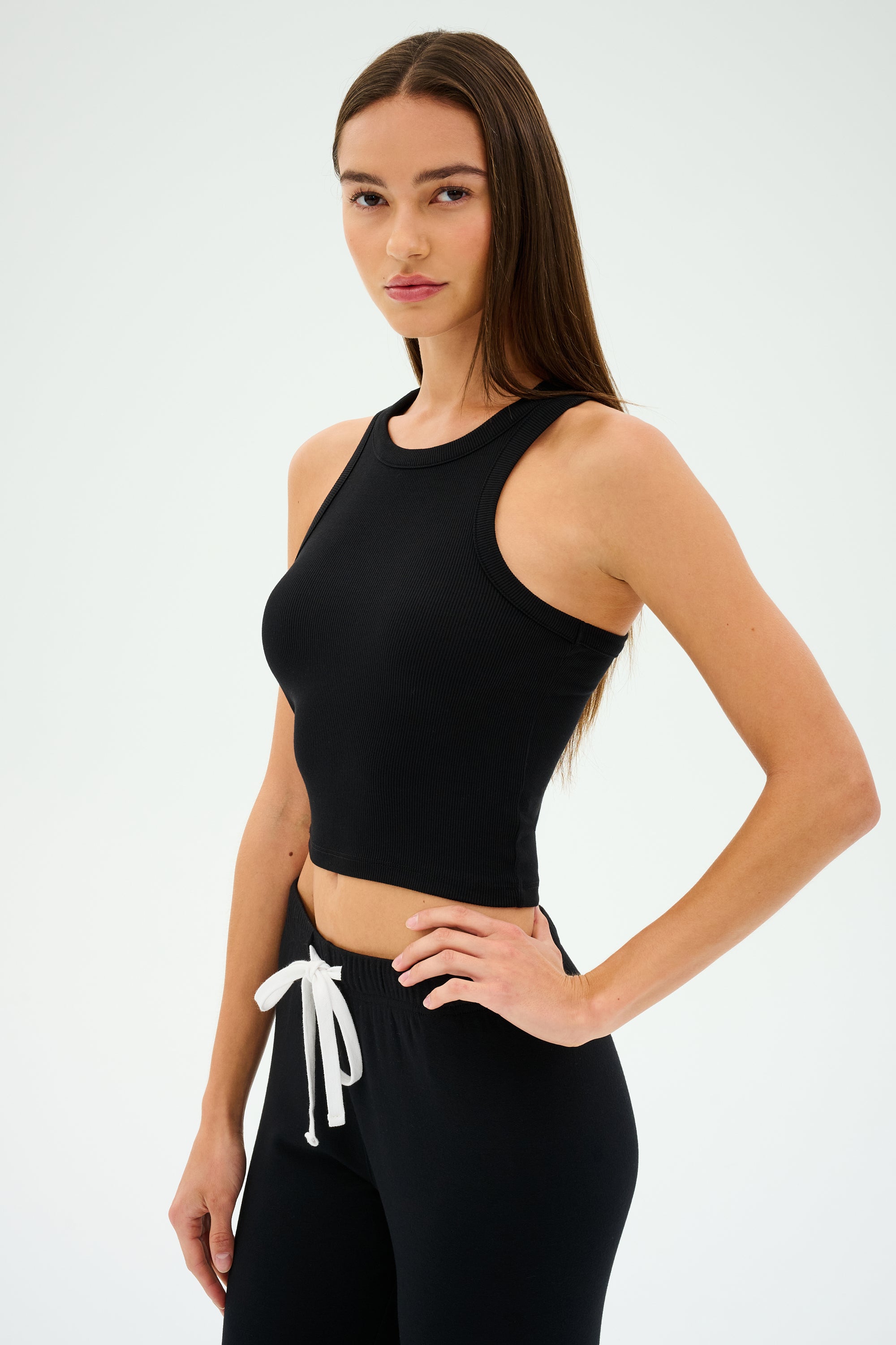 A person with long hair poses against a plain background wearing the SPLITS59 Kiki Rib Crop Tank in black, paired with black pants featuring a white drawstring.