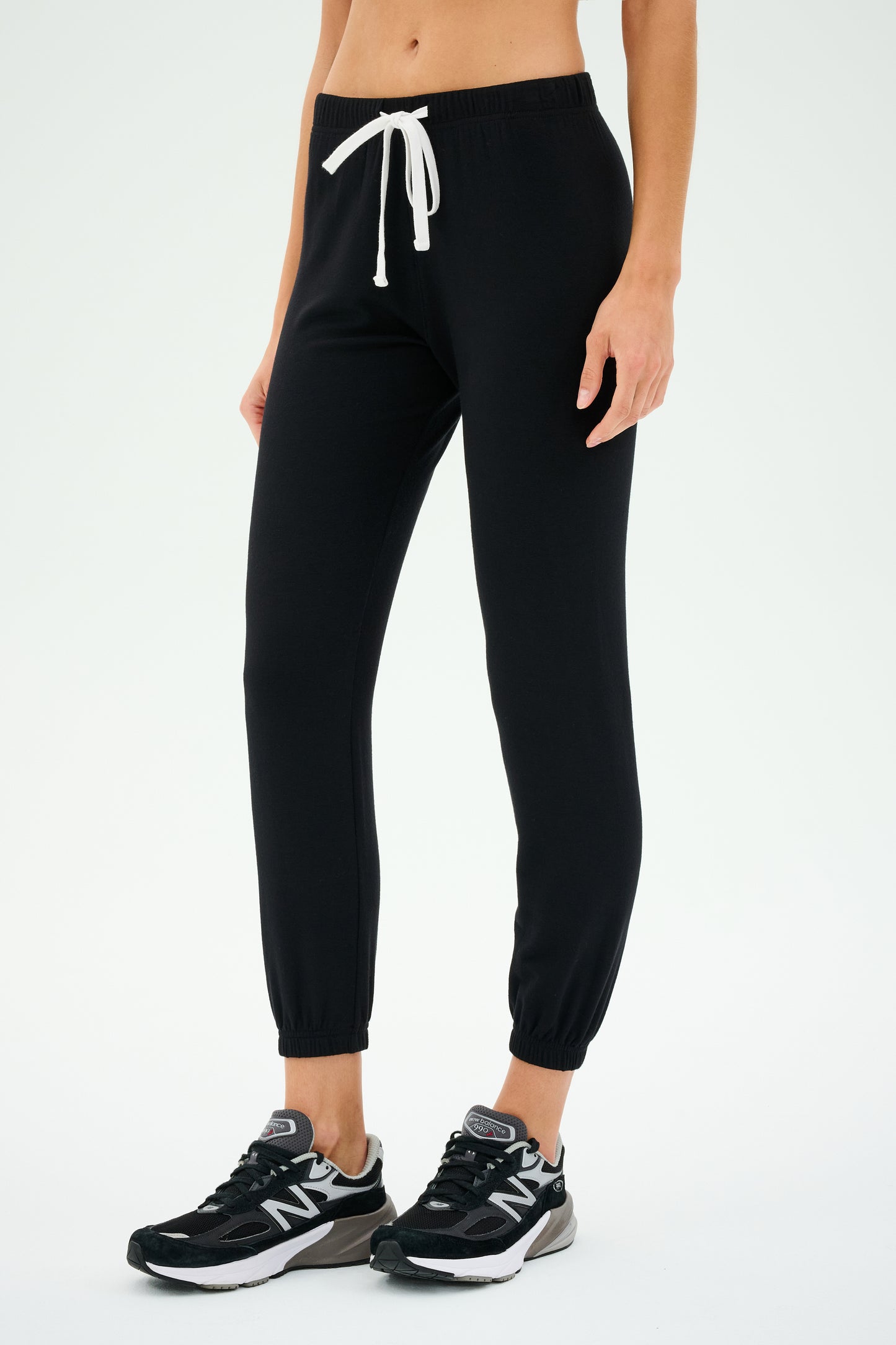 A person wearing the SPLITS59 Sonja Fleece Sweatpant - Black with a white drawstring and black New Balance sneakers stands against a plain background, ready for workouts.