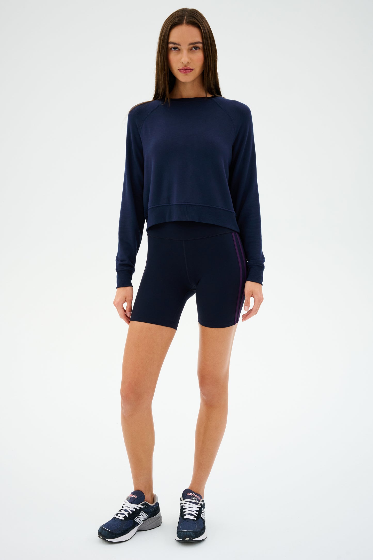 A person in a navy SPLITS59 Ella High Waist Airweight Short in Indigo/Black Currant, wearing sneakers, stands against a plain background.