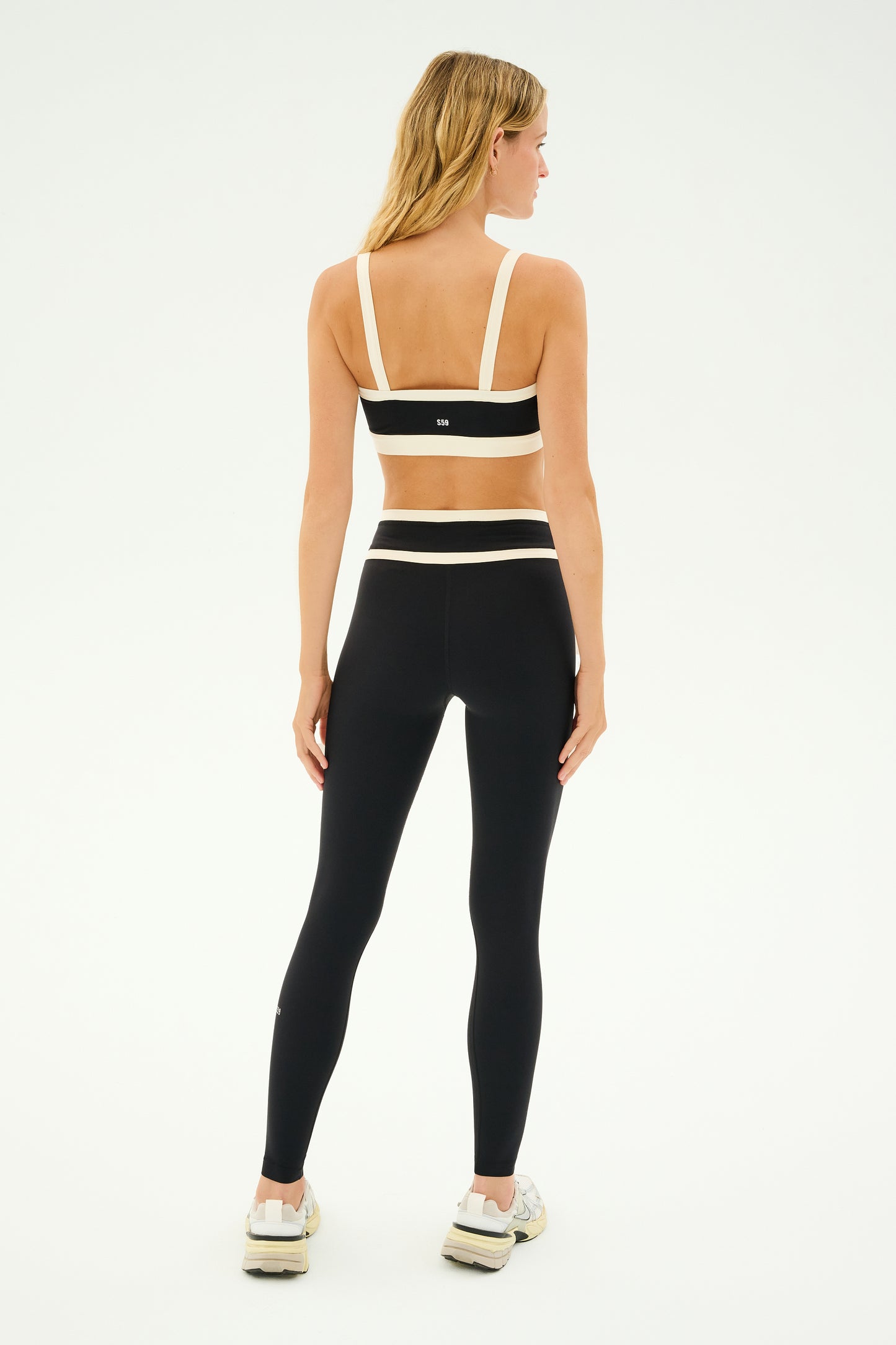 A person dressed in a black and white workout outfit, showcasing the SPLITS59 Monah Rigor Bra in Black/Creme, paired with sneakers, stands with their back turned.