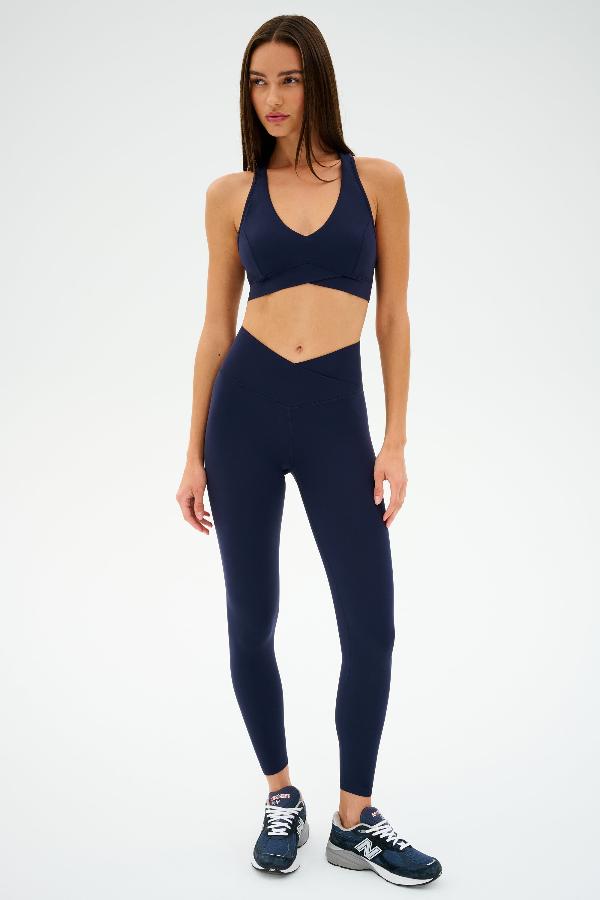 A person wearing a SPLITS59 Mia Rigor 7/8 navy sports bra and leggings, paired with navy sneakers, stands against a plain background.