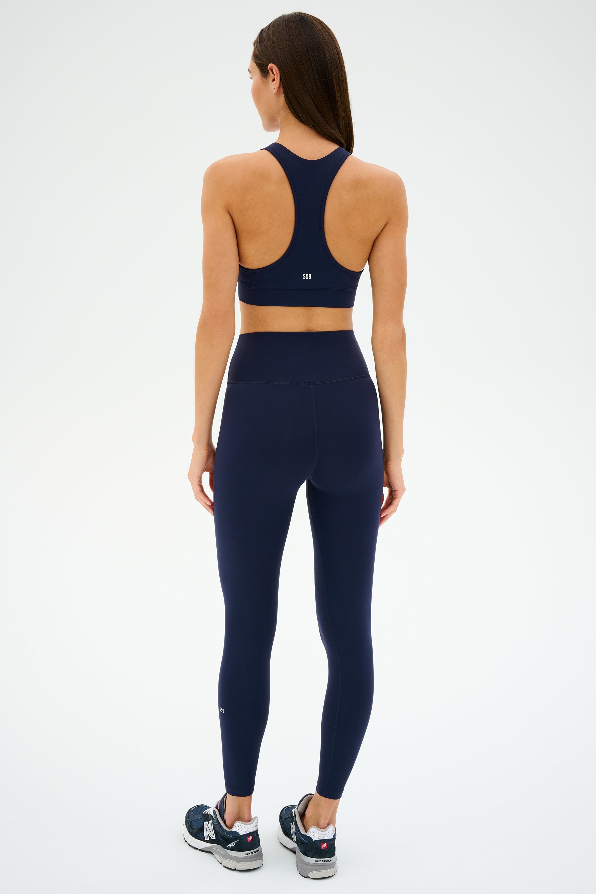 A person in a dark blue SPLITS59 outfit and Mia Rigor 7/8 Indigo leggings stands facing away from the camera, Rigor fabric sneakers completing their look against a plain background.