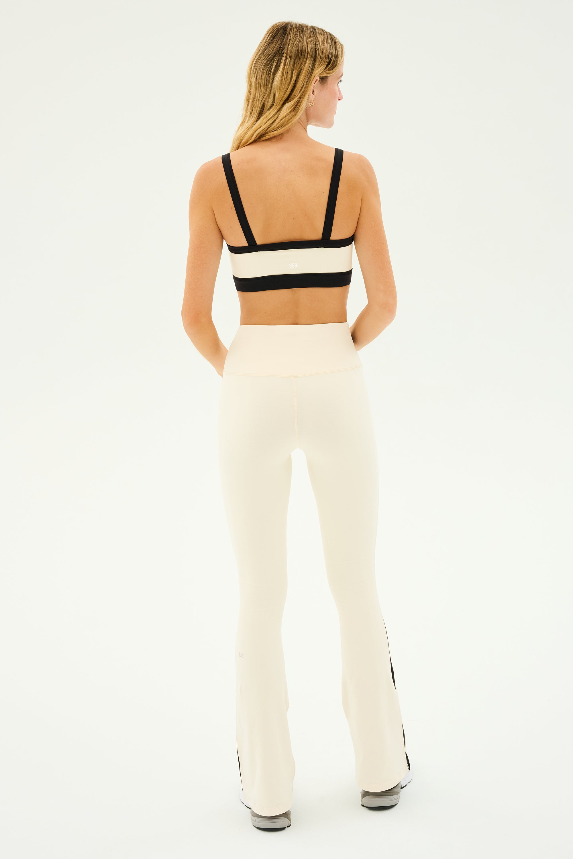 A person in a black and white sports outfit stands facing away, wearing the SPLITS59 Raquel High Waist Flared Legging in Creme/Black, paired with a supportive sports bra ideal for any workout.