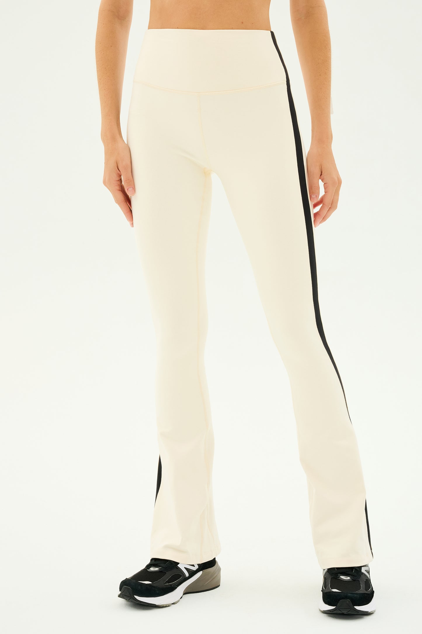 Against a plain background, an individual showcases the SPLITS59 Raquel High Waist Flared Legging in Creme/Black, featuring stylish black side stripes. Ideal for yoga workouts, these leggings offer a sleek design highlighted by 4-way stretch supplex for maximum comfort and flexibility, perfectly paired with black sneakers.