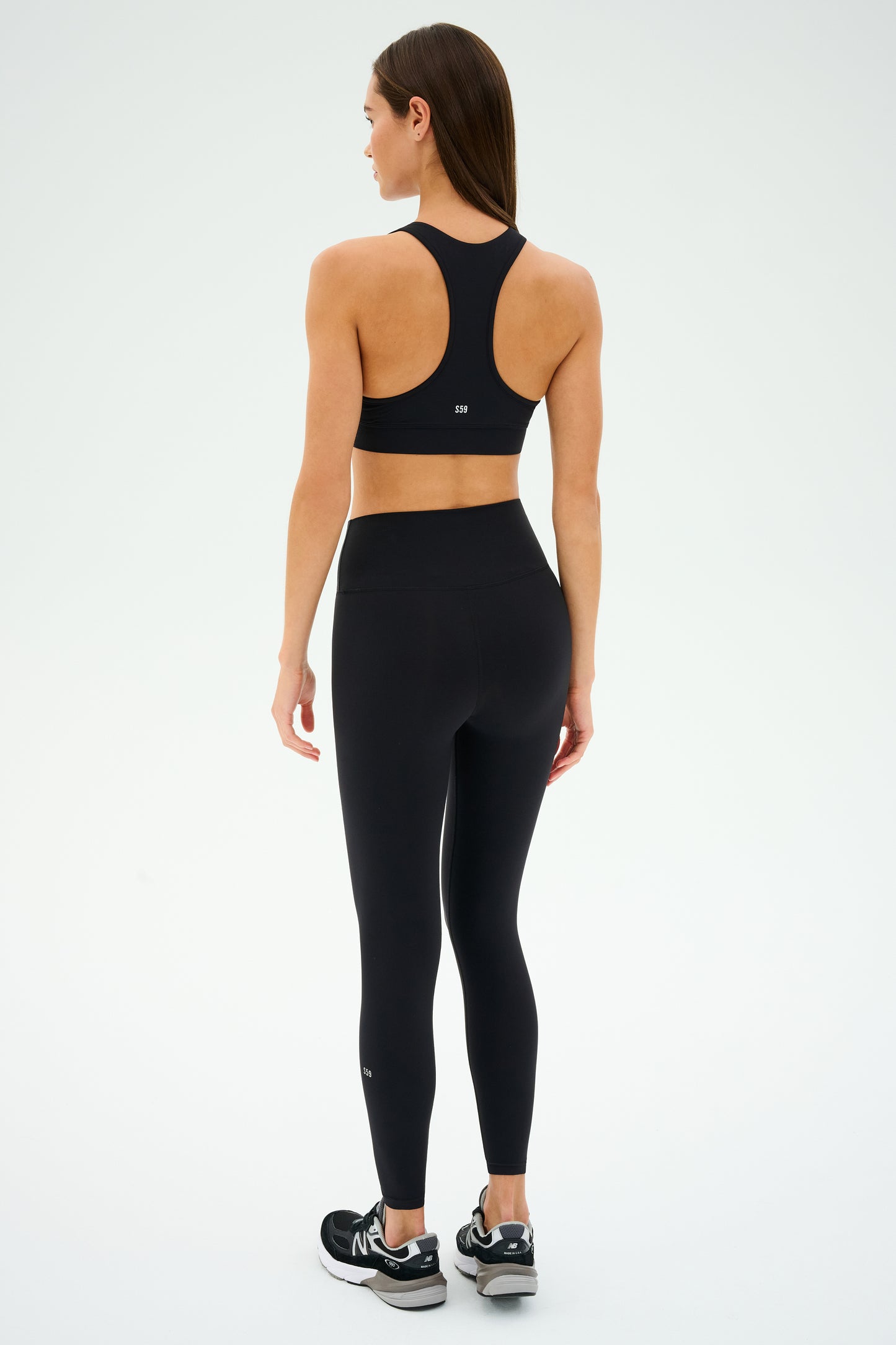 A person in Mia Rigor Bra - Black by SPLITS59, wearing black athletic wear and sneakers, showcases the ultra luxe Rigor fabric with their back to the camera against a white background.