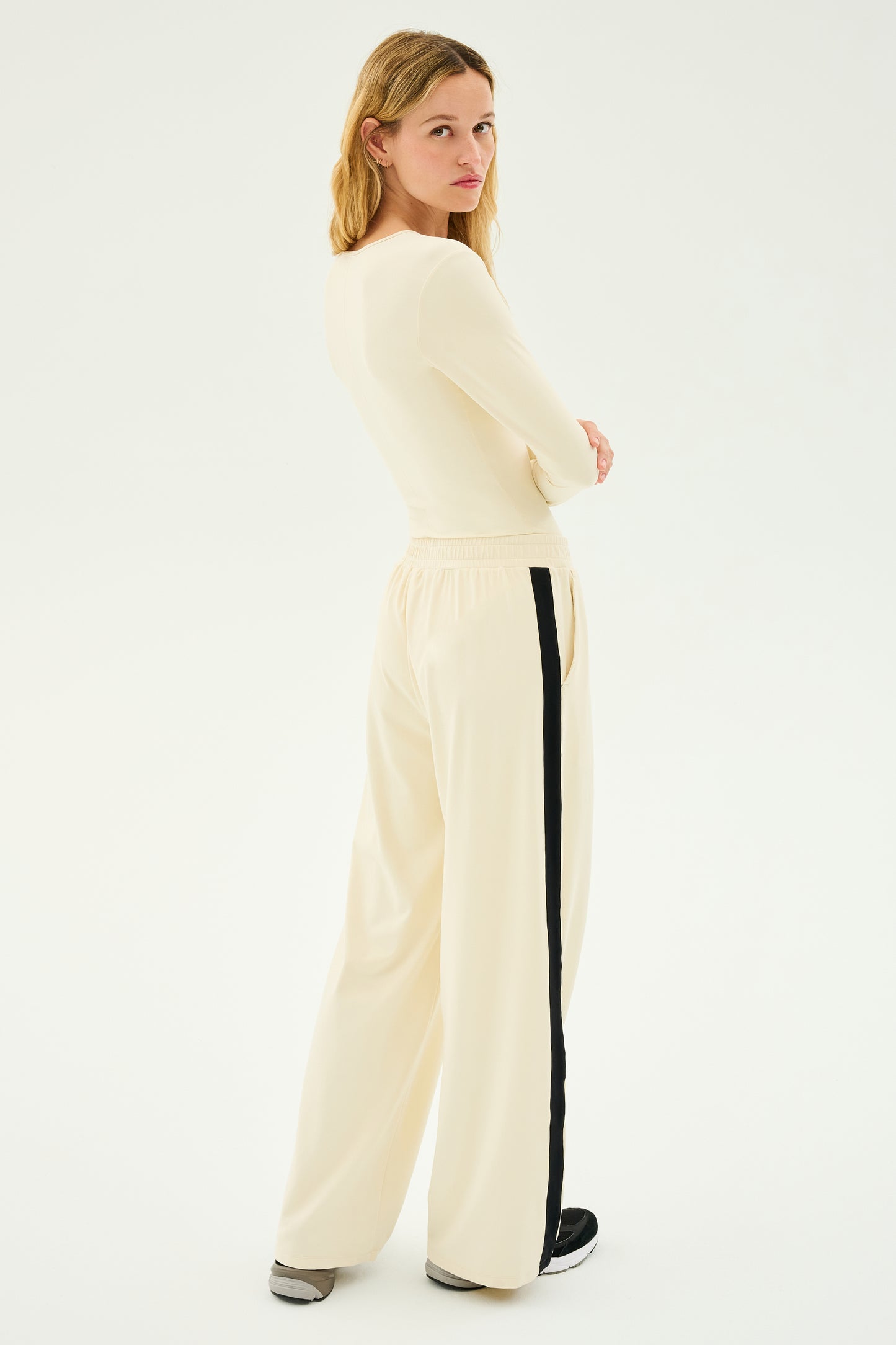 A woman facing slightly away, elegantly dressed in a cream long-sleeve top paired with SPLITS59's Luca Airweight Trouser with Stripe in Creme/Black, featuring wide-leg pants with sleek black stripes.