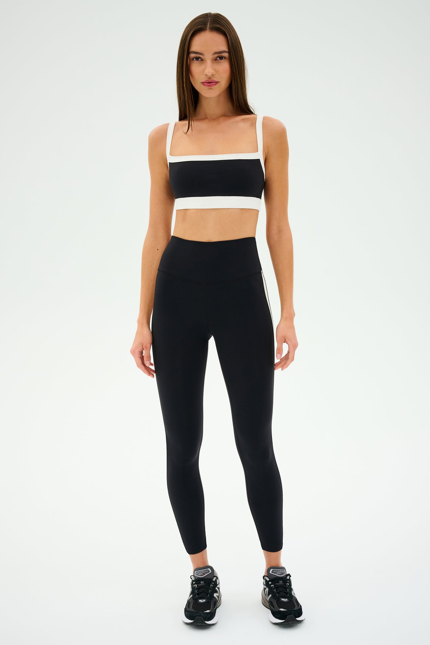 A person wearing a black and white sports outfit with black athletic shoes stands against a white background, channeling a retro sport vibe. The Monah Rigor Bra from SPLITS59 combines classic style with modern performance for solid support.