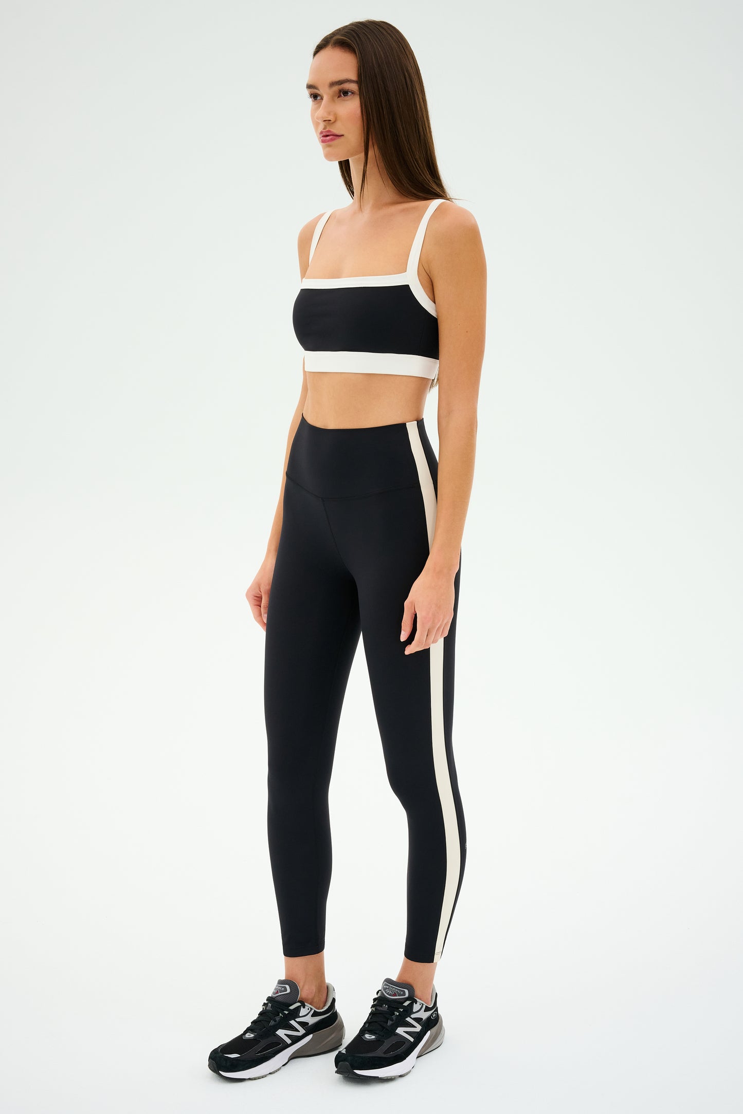 Dressed in a black and white athletic outfit, a woman confidently showcases the SPLITS59 Monah Rigor Bra for solid support, embodying a retro sport vibe against a plain background.