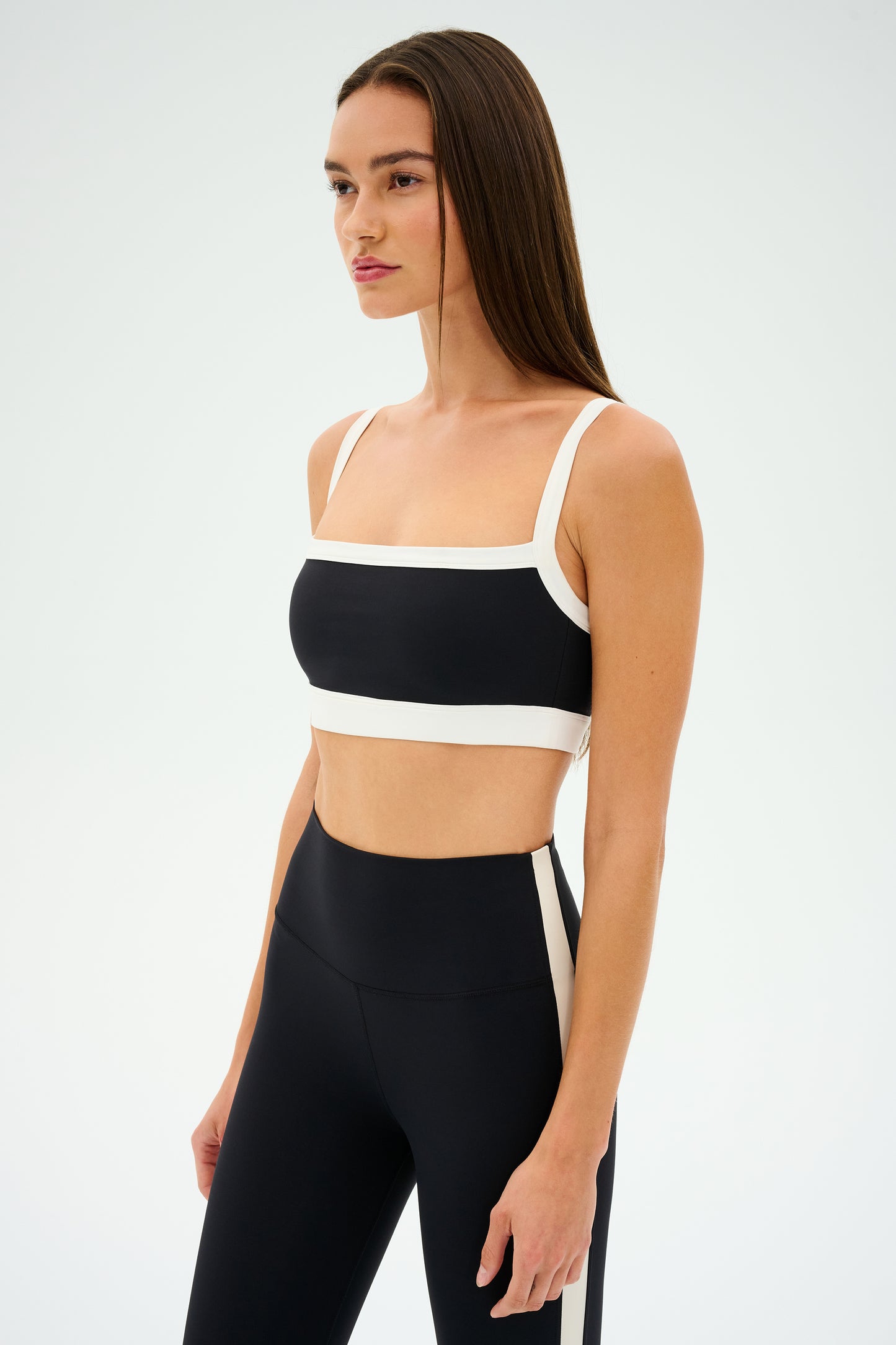 A woman in a black and white SPLITS59 Monah Rigor Bra stands against a plain background, facing left. The outfit seamlessly blends solid support with a retro sport vibe, creating a stylish yet functional appearance.