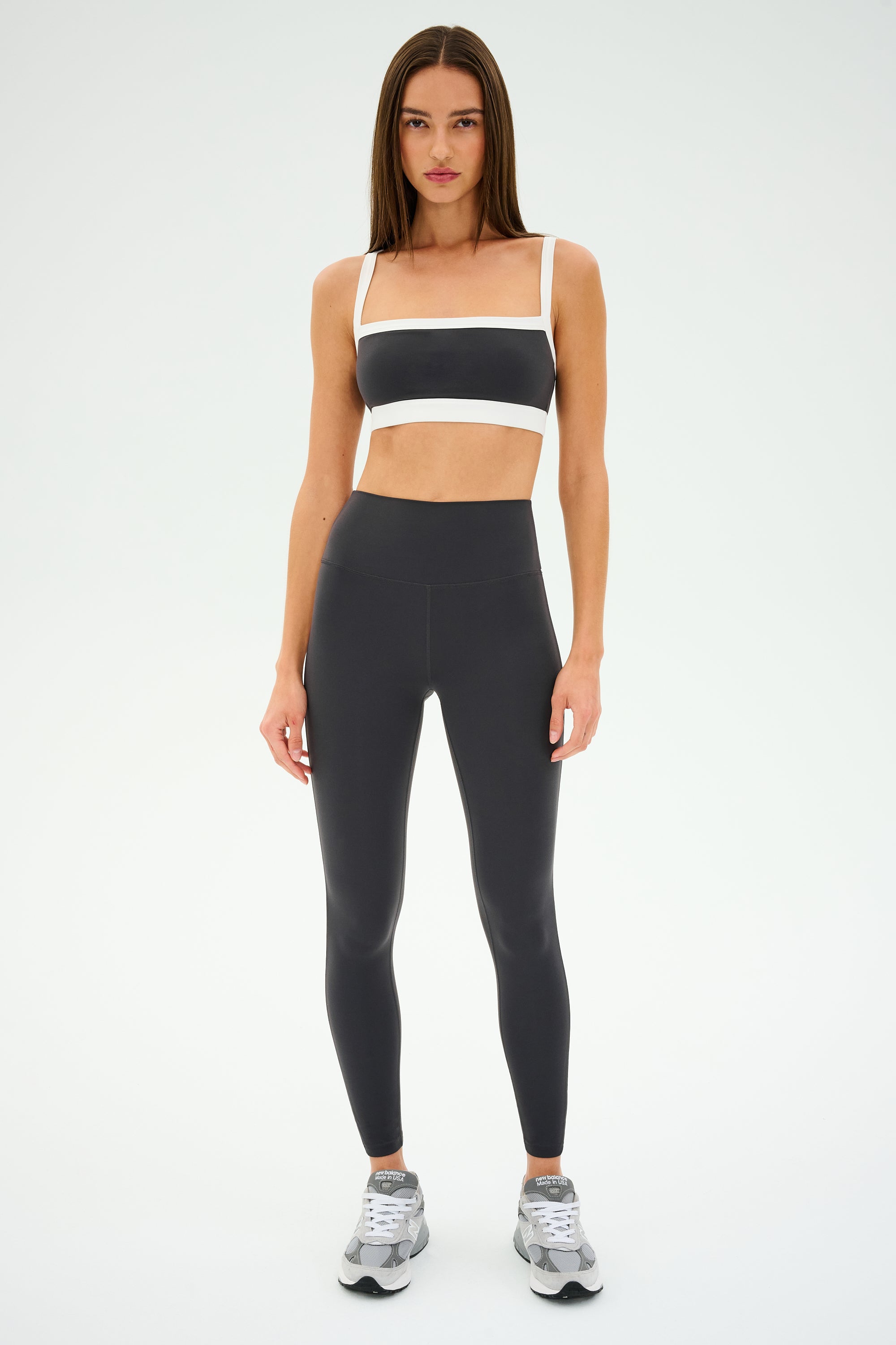 A person stands facing forward, wearing a black sports bra with white straps and SPLITS59 Clare High Waist Rigor 7/8 leggings in Graphite/White, ideal for gym workouts. Gray sneakers complete the look against a plain light background, highlighting the high-performance Rigor fabric's sleek design.