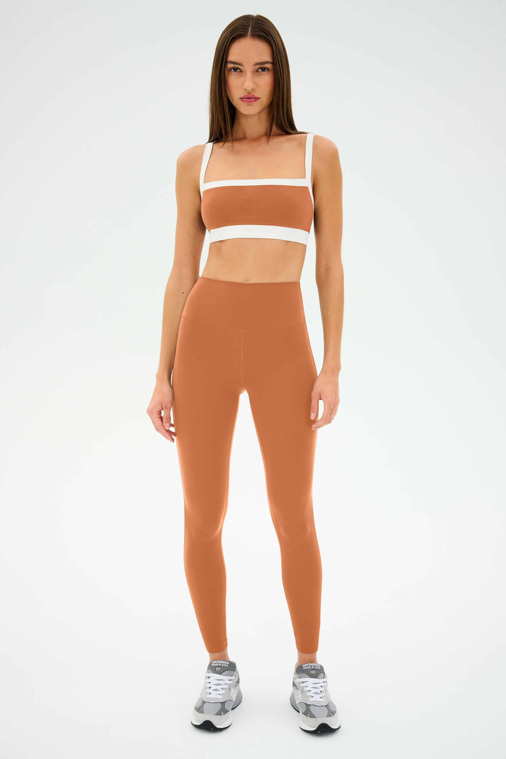 A person models SPLITS59's Monah Rigor Bra in pecan/white, paired with matching leggings and gray sneakers, on a plain white background.