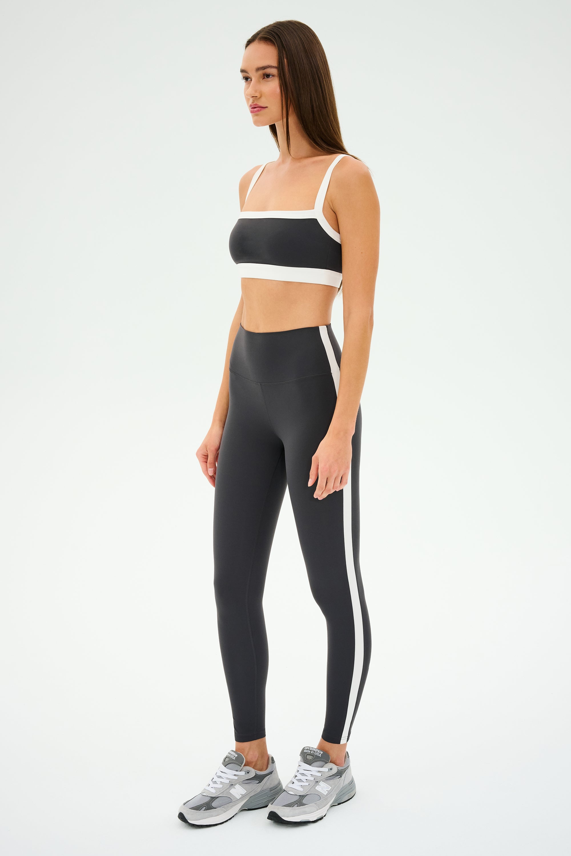 Someone wearing a black and white sports outfit featuring SPLITS59's Clare High Waist Rigor 7/8 leggings in Graphite/White and silver sneakers, ideal for gym workouts, stands confidently on a white background.