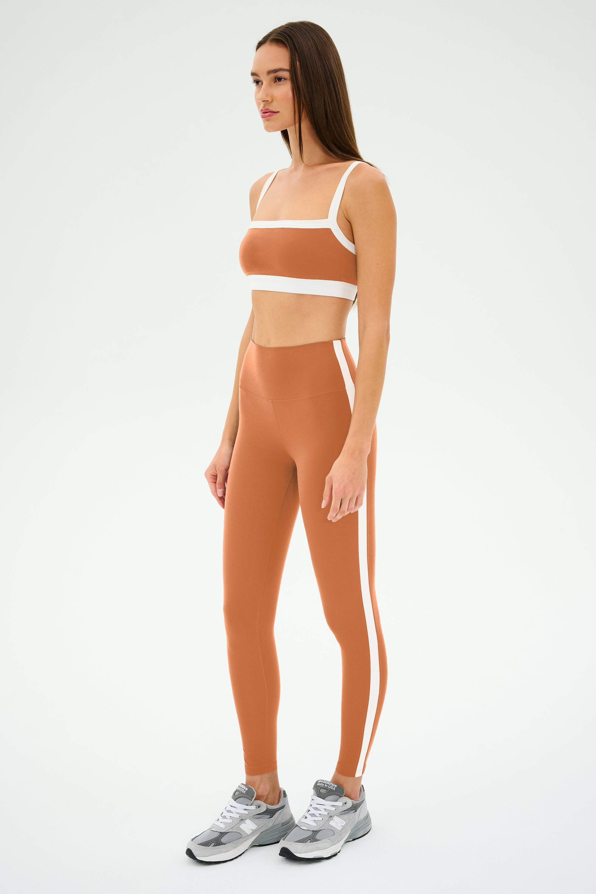 A woman poses left in SPLITS59's Clare High Waist Rigor 7/8 in Pecan/White with a nylon-spandex crop top and silver sneakers against a plain backdrop.