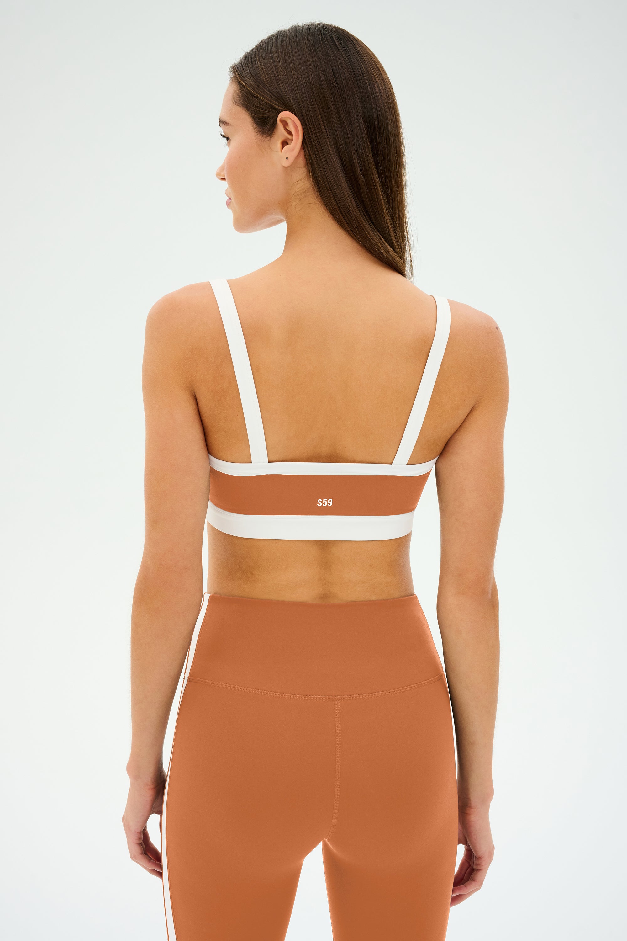 A woman with long hair is wearing the SPLITS59 Monah Rigor Bra in Pecan/White and matching leggings made from breathable spandex, viewed from the back.