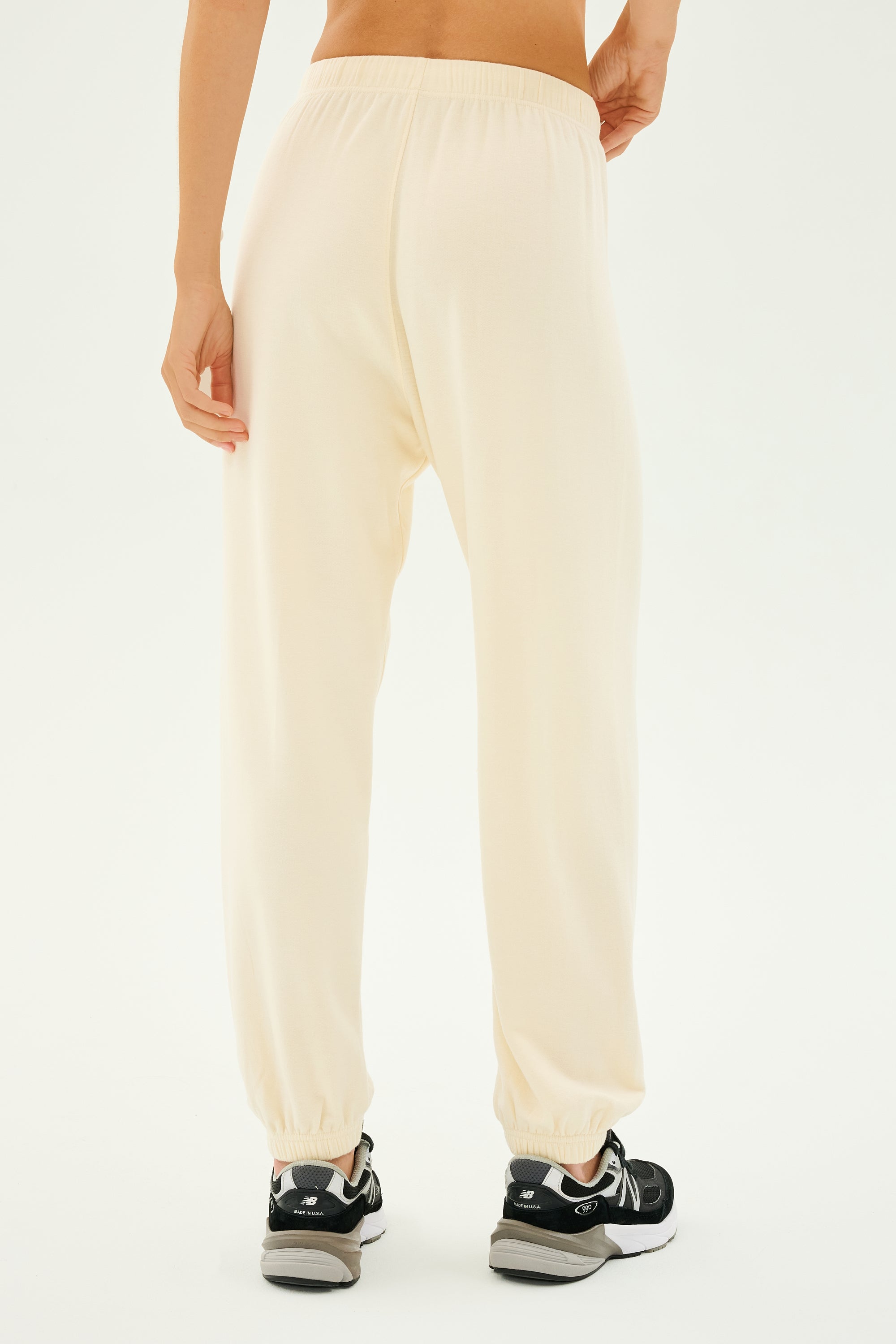 A person stands against a plain background, wearing the Andie Oversized Fleece Sweatpants in Creme by SPLITS59, perfectly suited for workouts and effortlessly blending comfort with a touch of flair alongside stylish black sneakers.