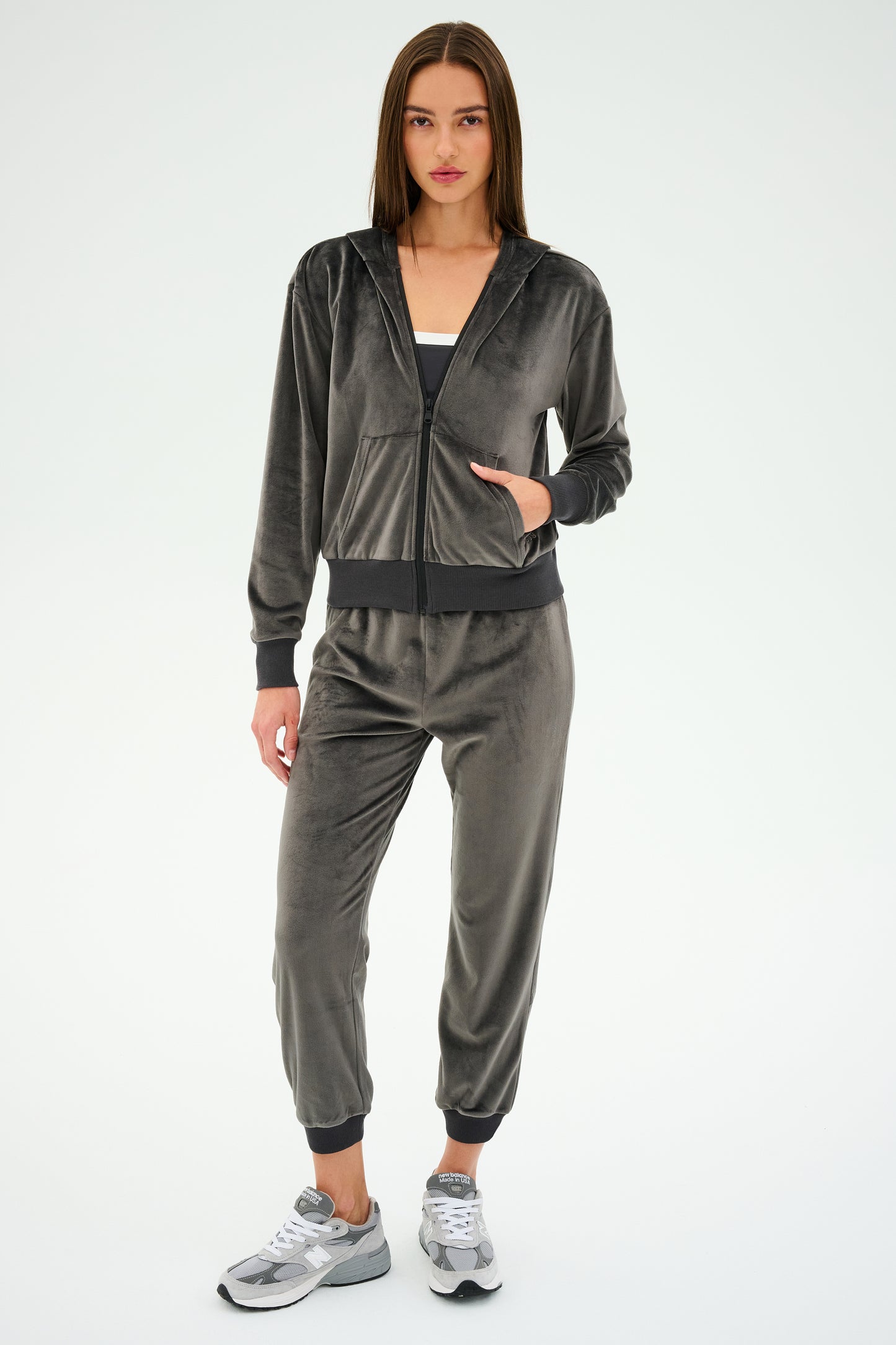 Wearing SPLITS59's Rhys Velour Jogger in Graphite, a person pairs the luxe velour tracksuit with a white top and silver sneakers against a plain white backdrop.