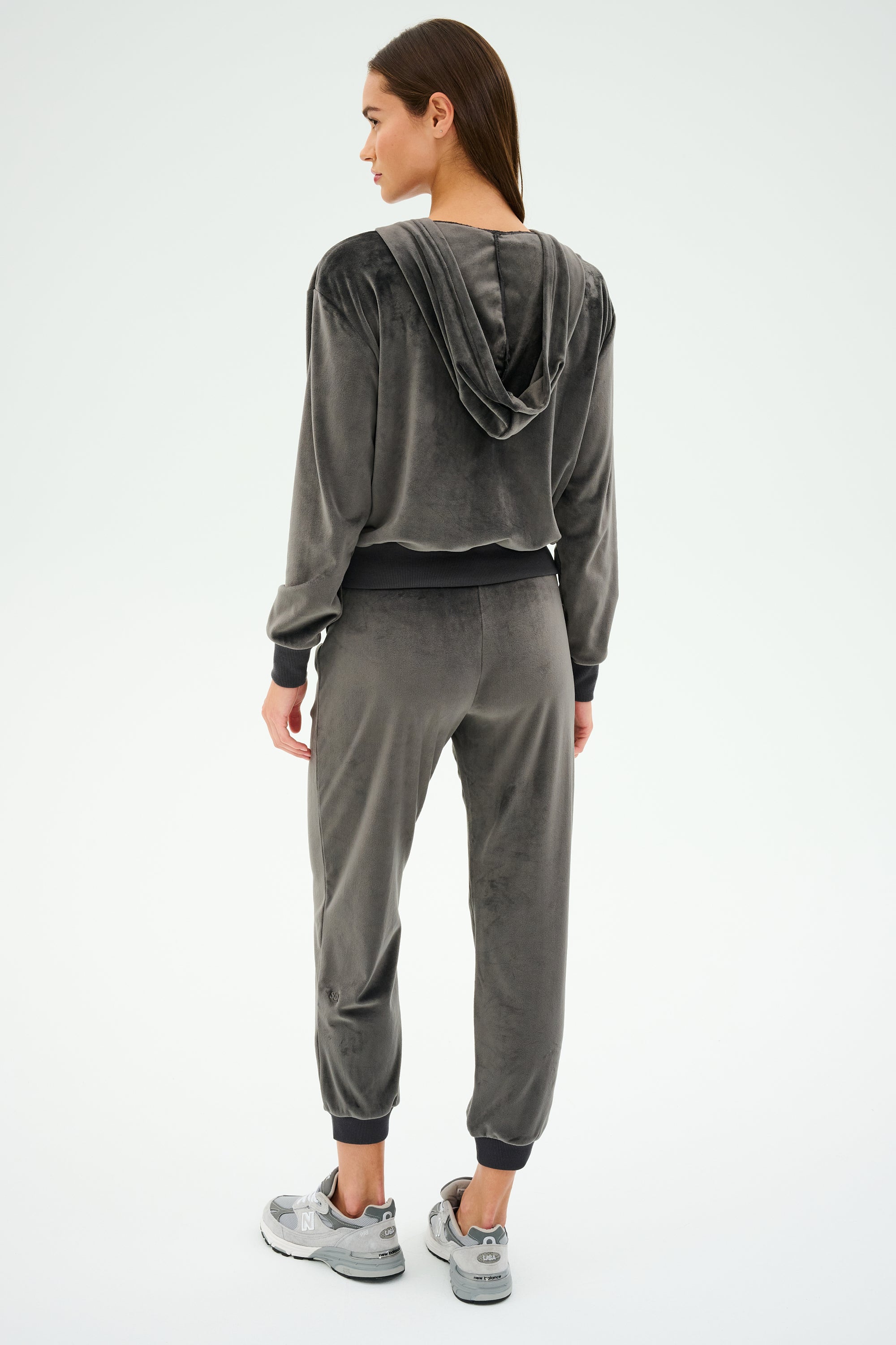 Wearing SPLITS59's Rhys Velour Jogger in Graphite with sneakers, a person stands to the side, highlighting the luxurious velour fabric against a light backdrop.