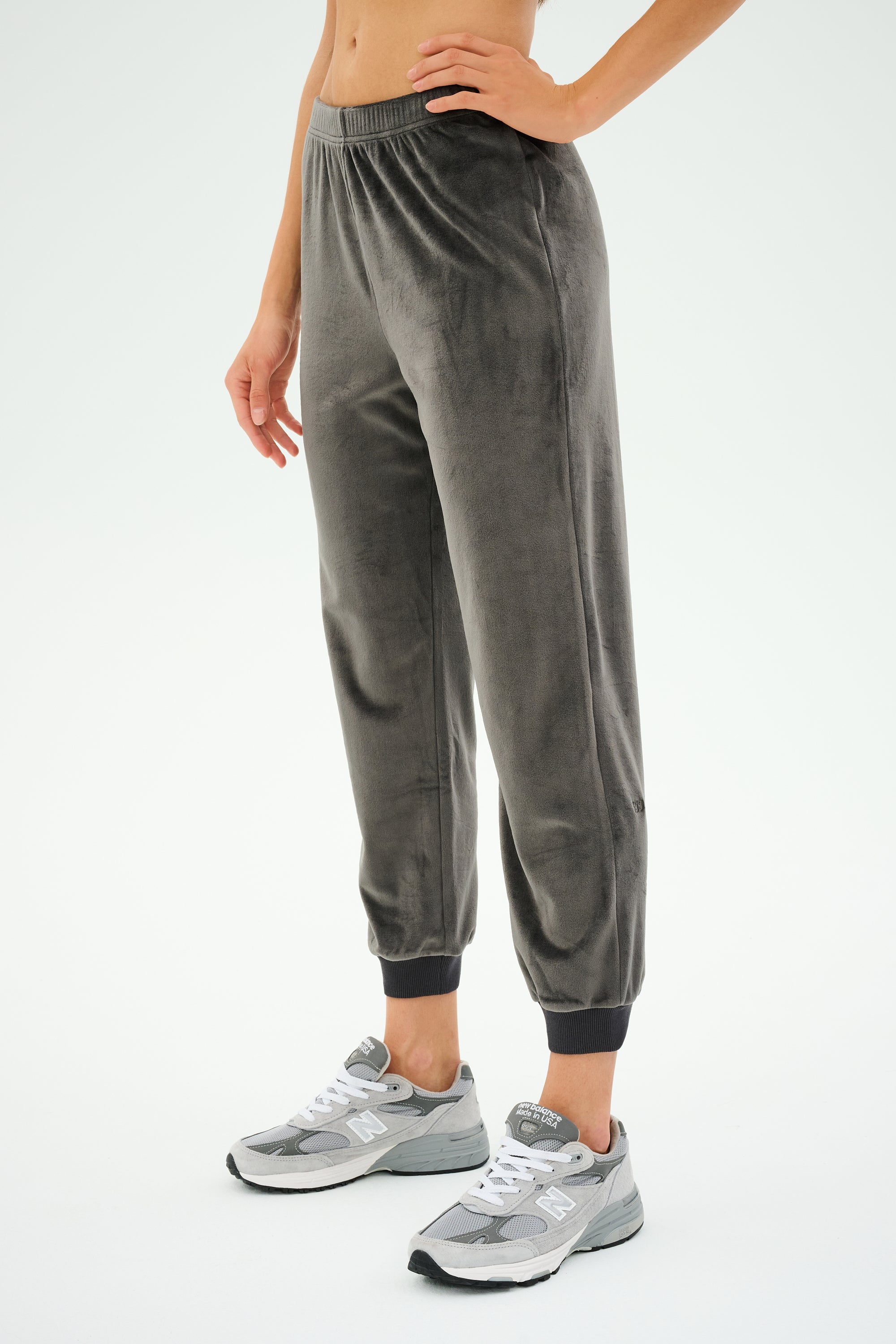 Dressed in casual attire, the person showcases the SPLITS59 Rhys Velour Jogger in Graphite, paired seamlessly with matching gray sneakers against a plain background. The luxurious velour fabric adds an elegant touch to this relaxed outfit.