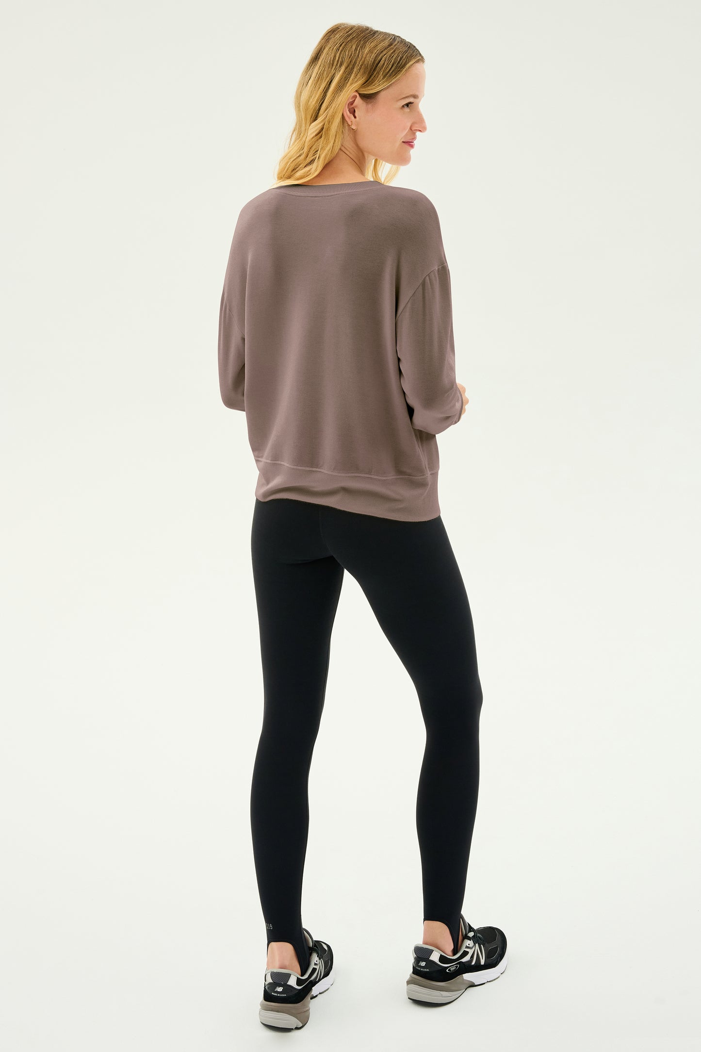 Sporting shoulder-length hair, a person is wearing the Sonja Fleece Sweatshirt in Lentil by SPLITS59, paired with black leggings and black sneakers. They are standing against a plain background, facing slightly away. The MADE IN LOS ANGELES style flawlessly combines comfort and sophistication in their look.