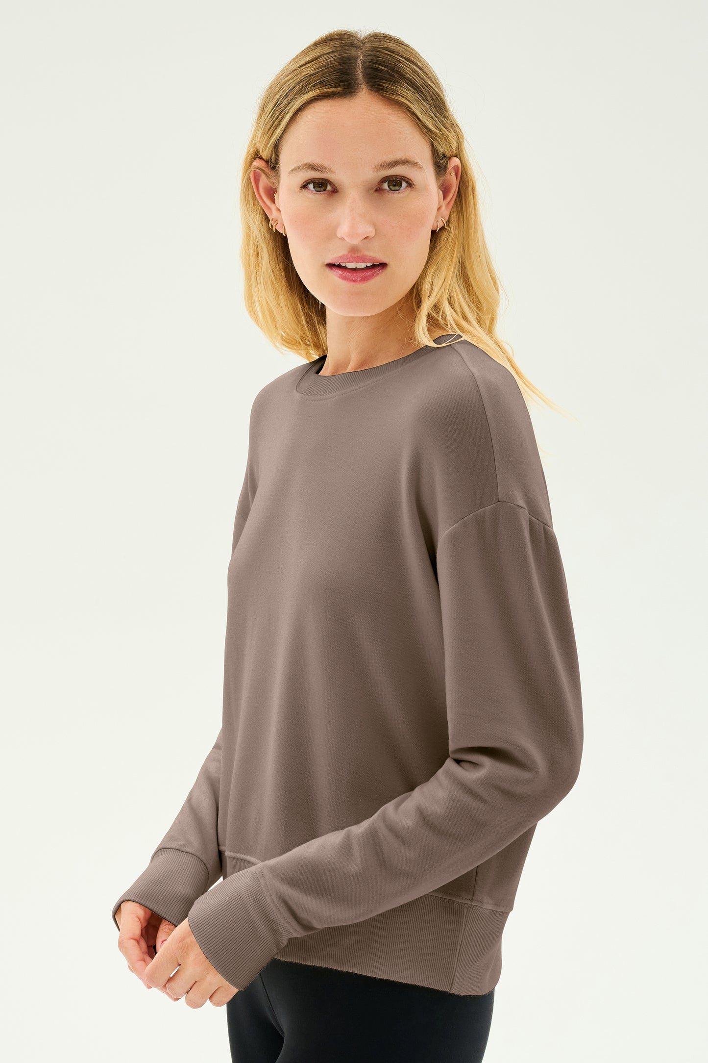 A person with long blonde hair stands against a plain light background, wearing the Sonja Fleece Sweatshirt in Lentil by SPLITS59 and black pants.