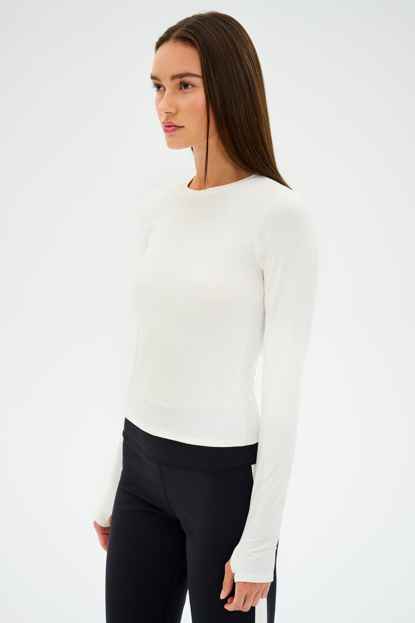 A woman in an Airweight Lite LS Waist Length from SPLITS59 and black pants stands against a plain background, looking to the side.