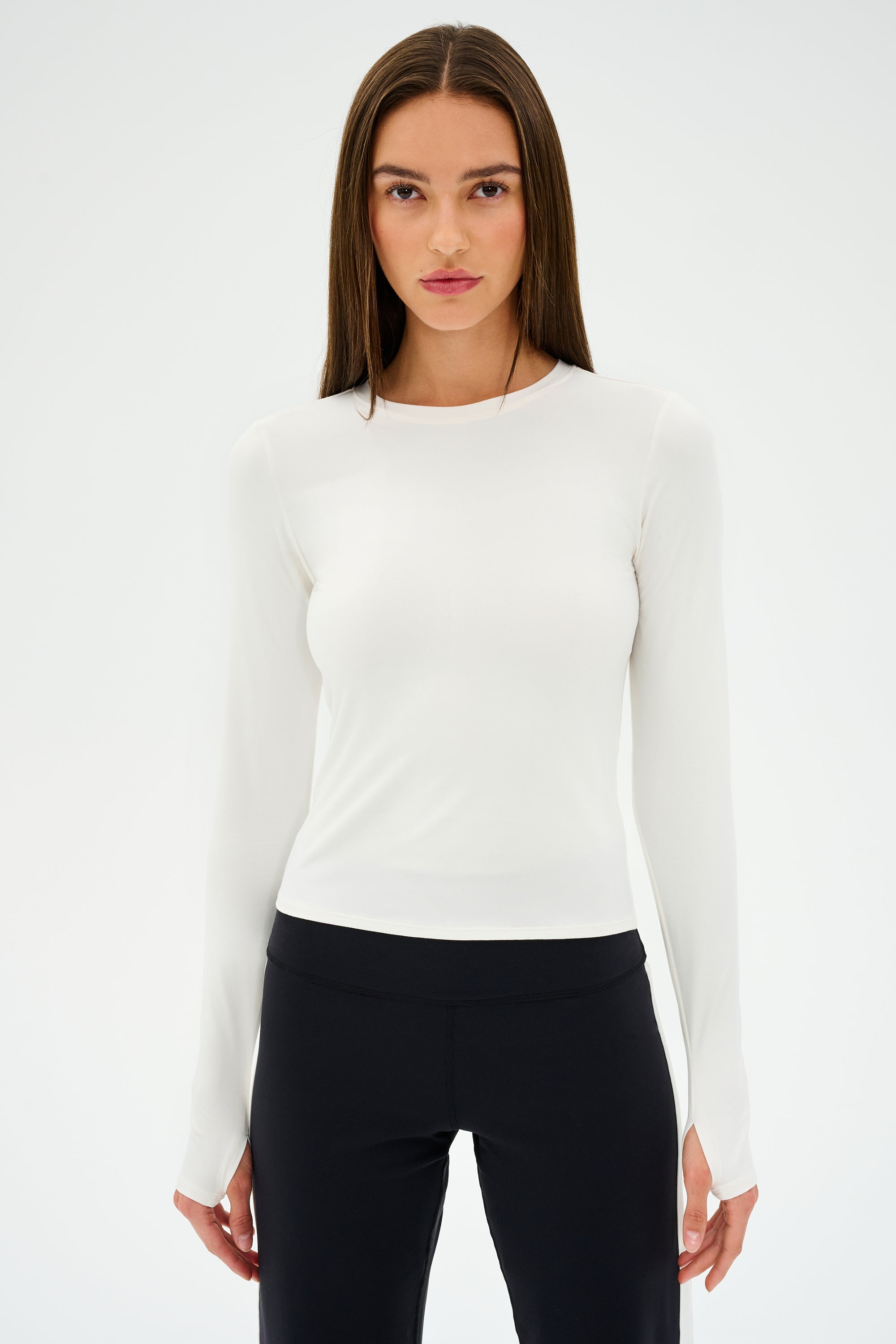 A person with long hair is wearing a white SPLITS59 Airweight Lite LS Waist Length tee and black pants, standing against a plain background.