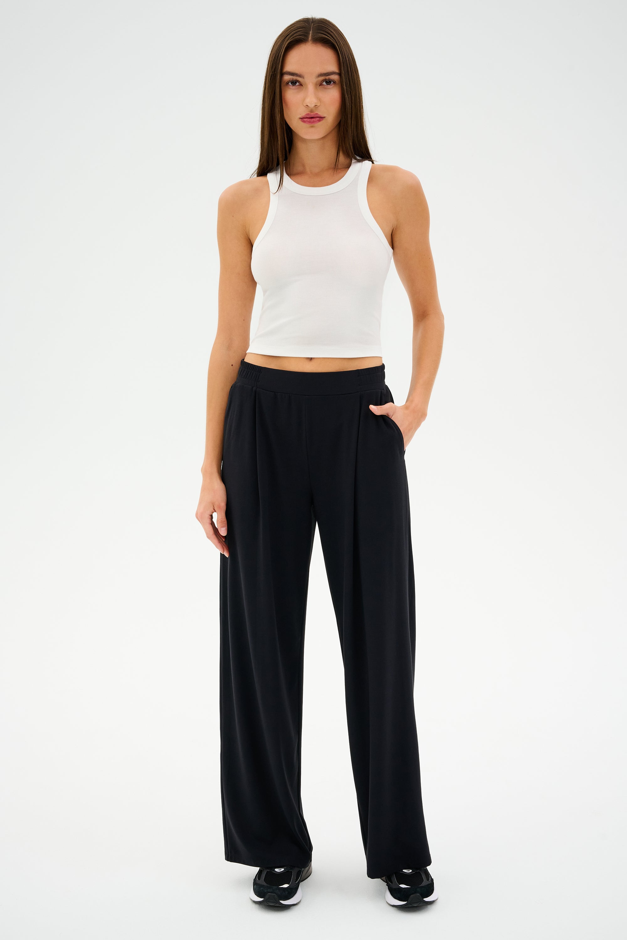 Standing against a plain backdrop, someone showcases style and comfort with a white sleeveless top, black SPLITS59 Lucas Airweight Trousers, coordinated shoes, and one hand resting casually in a pocket.