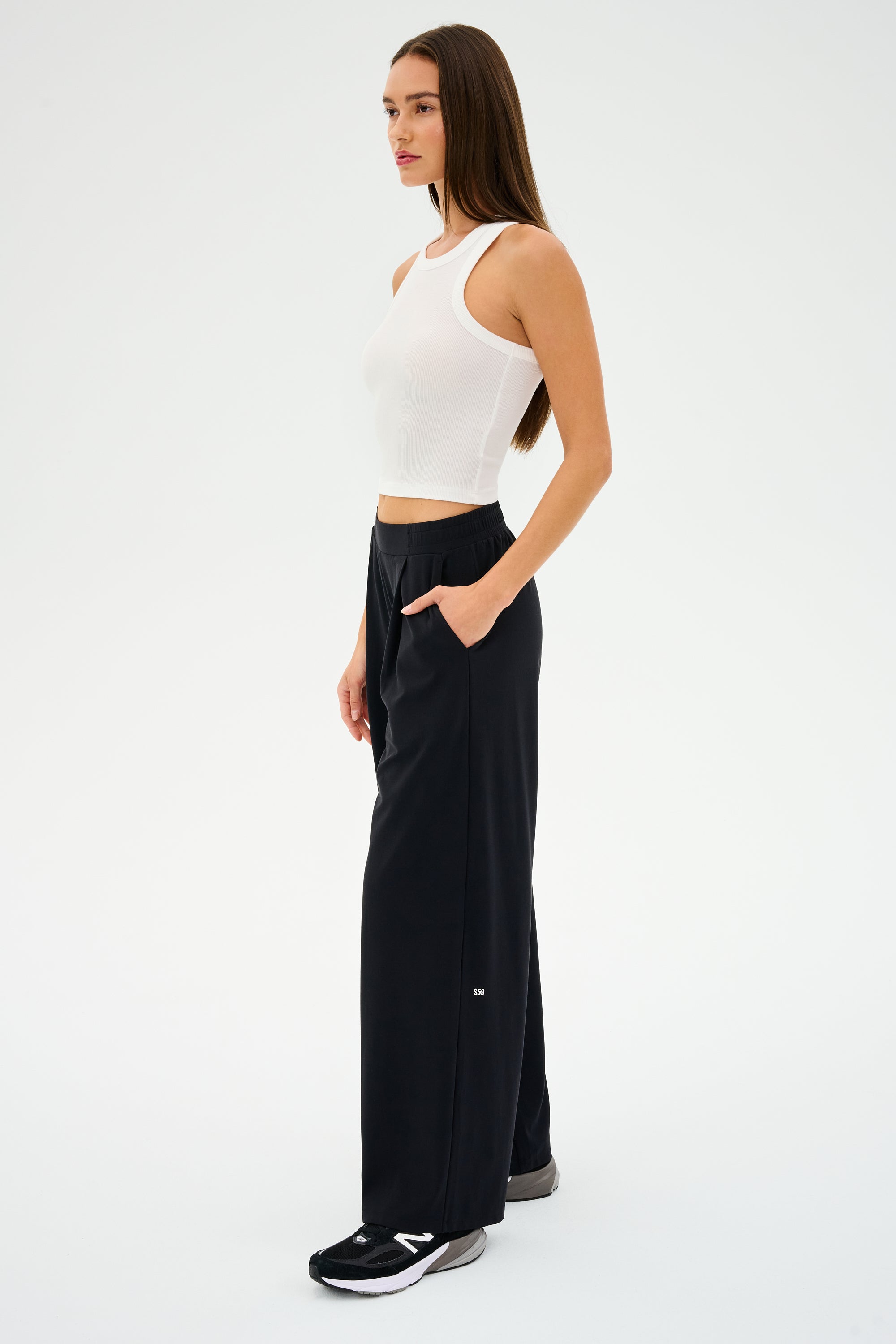 A person in a white sleeveless top and SPLITS59's Lucas Airweight Trouser - Black, made from ultra-luxe fabric, stands in profile with hands in pockets, showcasing sophisticated comfort against a plain background.