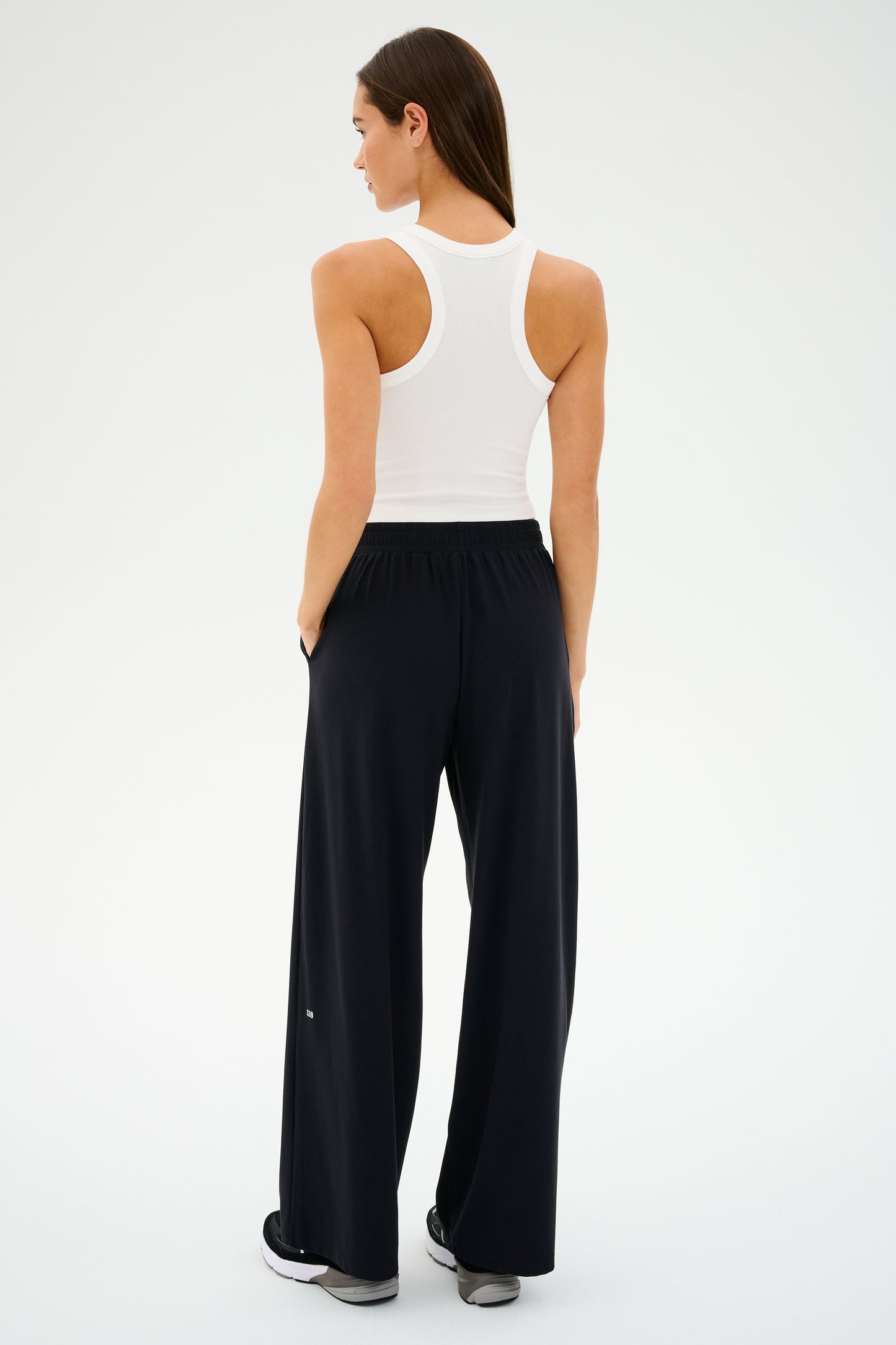 A person in a white tank top and black SPLITS59 Lucas Airweight Trouser stands with their back to the camera, radiating sophisticated comfort in a plain setting.