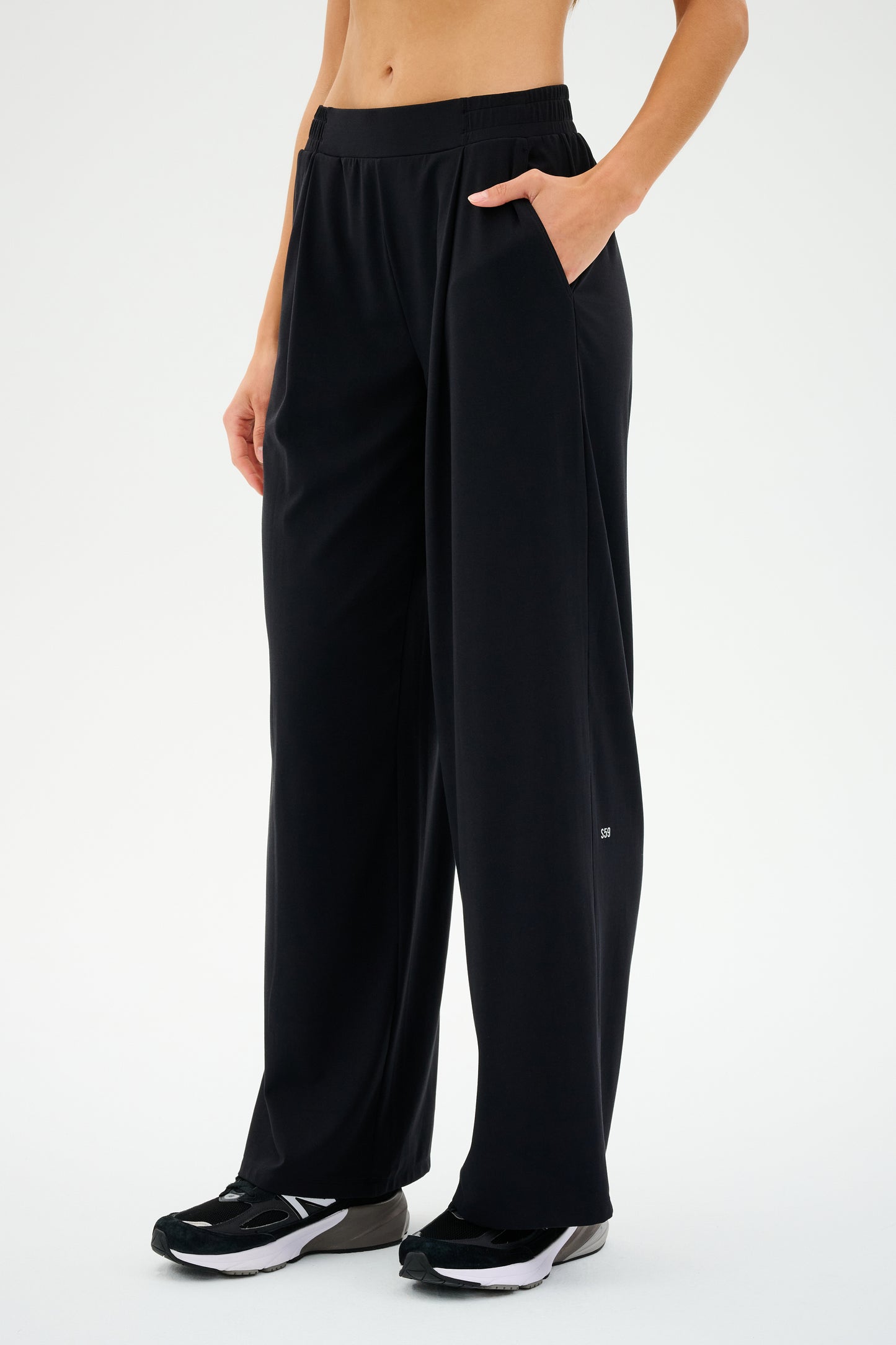 Clad in SPLITS59's Lucas Airweight Trouser in black, the individual exudes effortless sophistication. Posing confidently against a white backdrop, they wear stylish black sneakers with one hand casually tucked into a pocket.