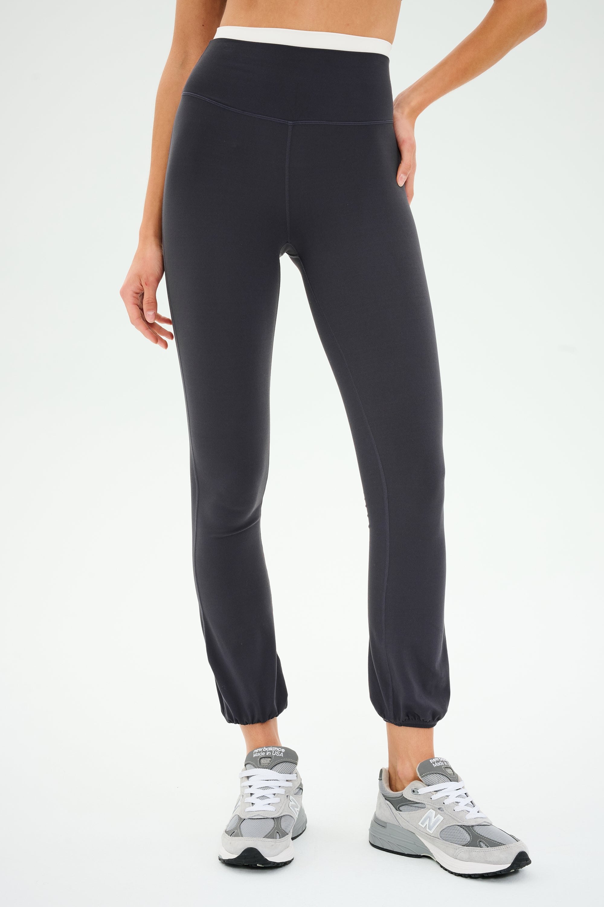 A person confidently stands with one hand on the hip, wearing SPLITS59's Icon Dual High Waist Airweight Legging in Graphite and gray sneakers against a plain background.