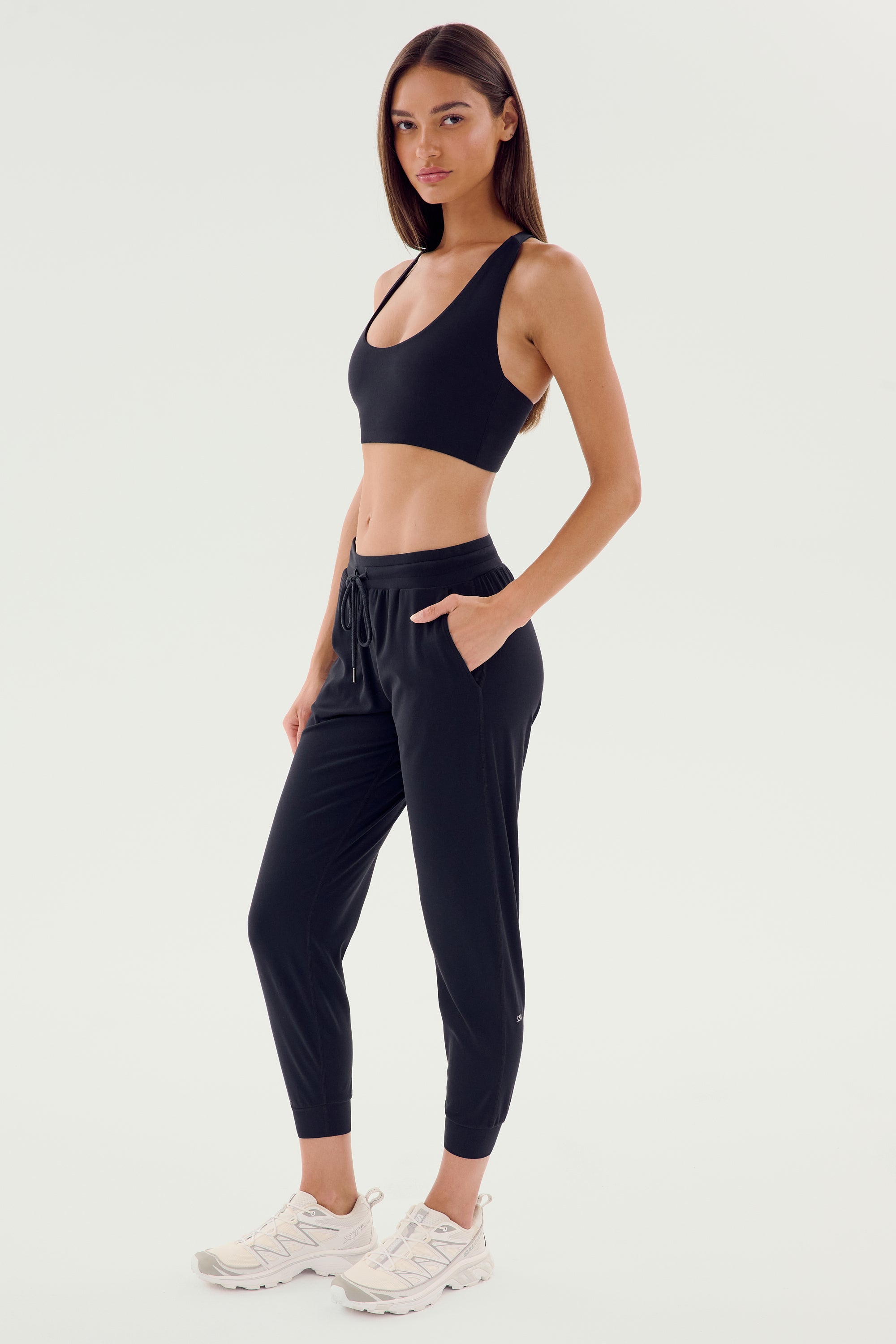 A person in Classic Airweight Joggers by SPLITS59, dressed in black workout wear made from Airweight fabric and white sneakers, strikes a pose against a plain background.