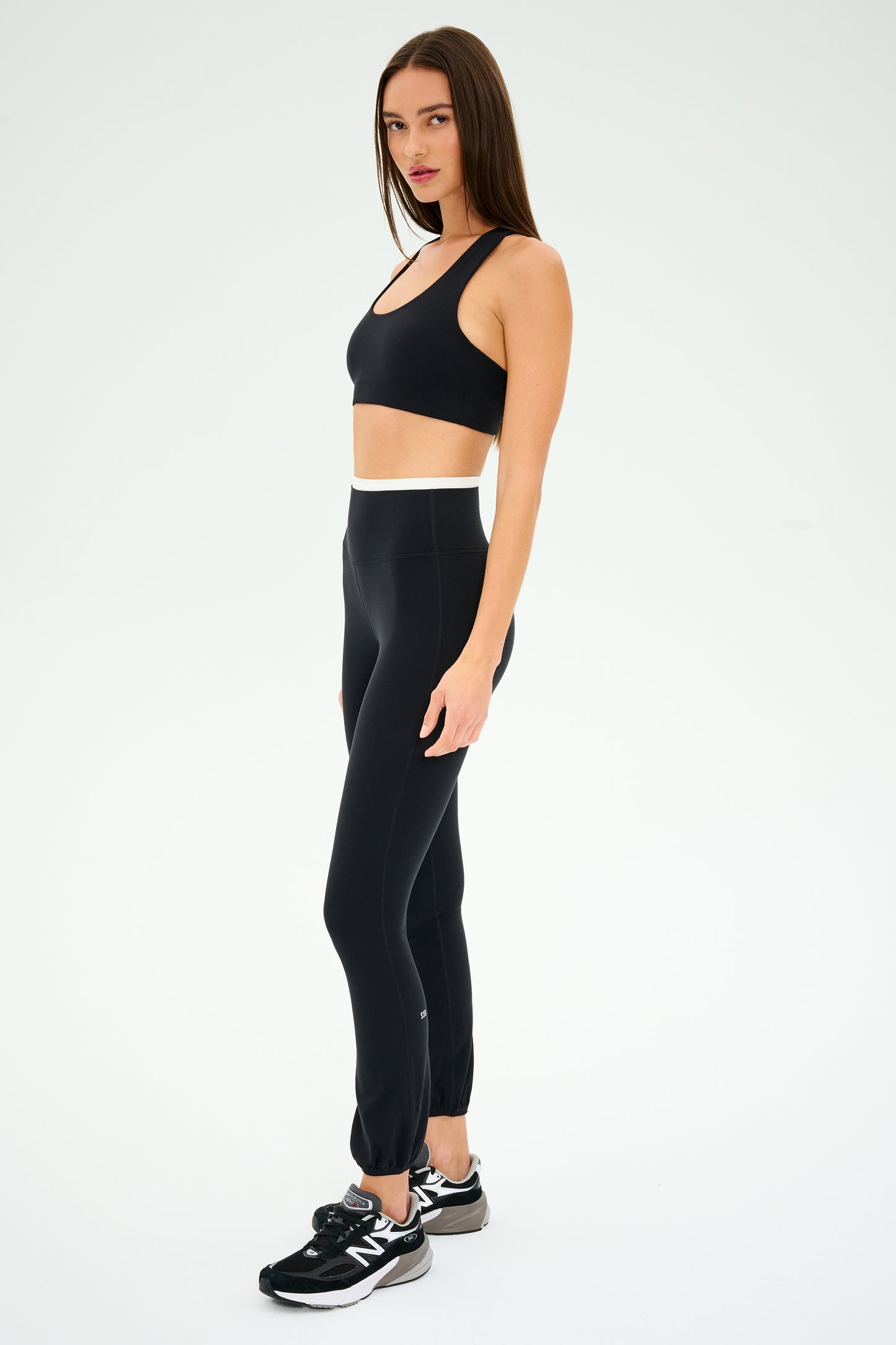 A person is wearing a black sports bra, SPLITS59 Icon Dual High Waist Airweight Leggings - Black, and black sneakers, standing against a plain white background.